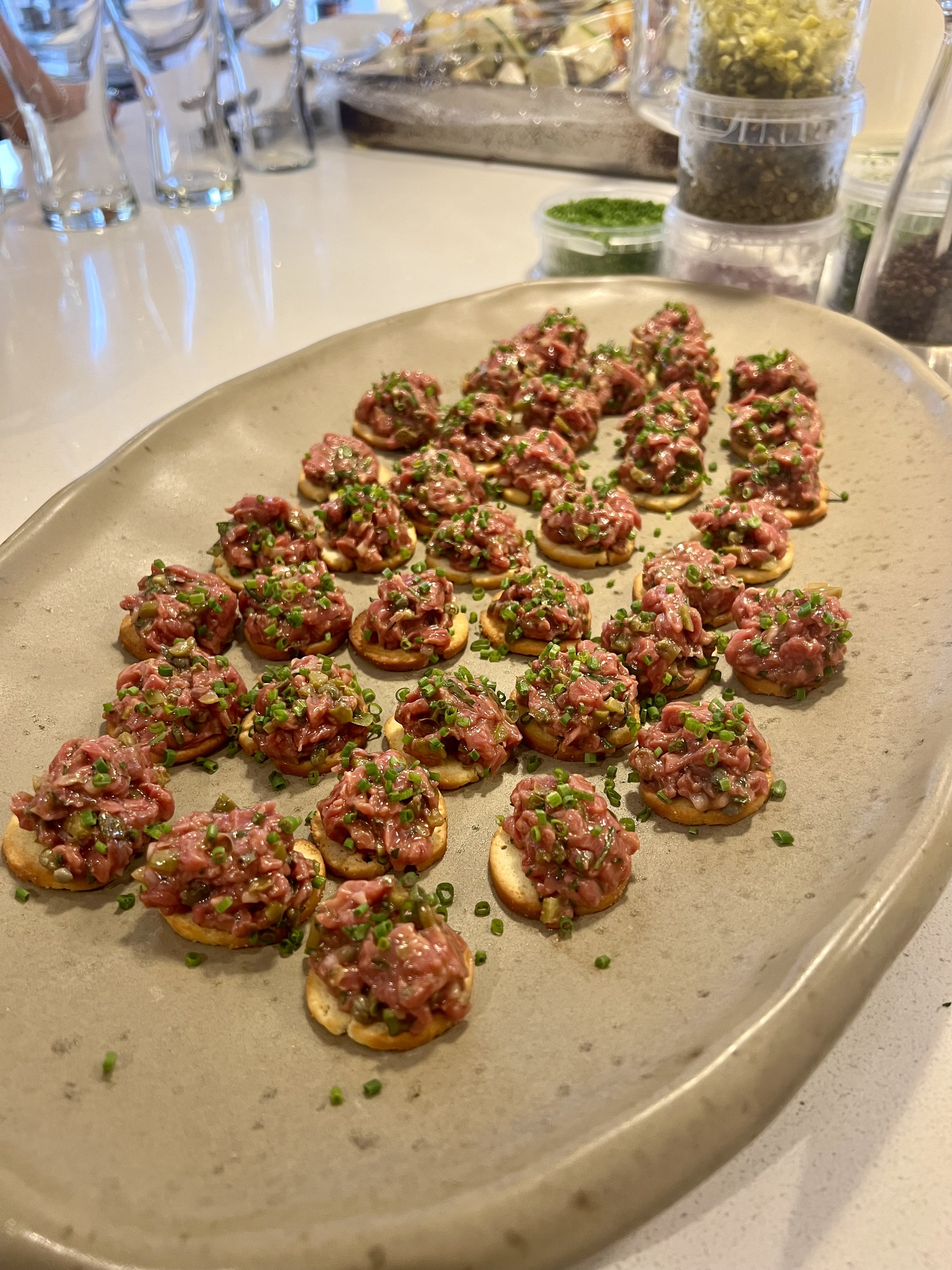 Close-up of a plate with small appetizers topped with a mixture of minced meat and chopped green herbs, garnished with chopped green onions, on a kitchen or dining table.
