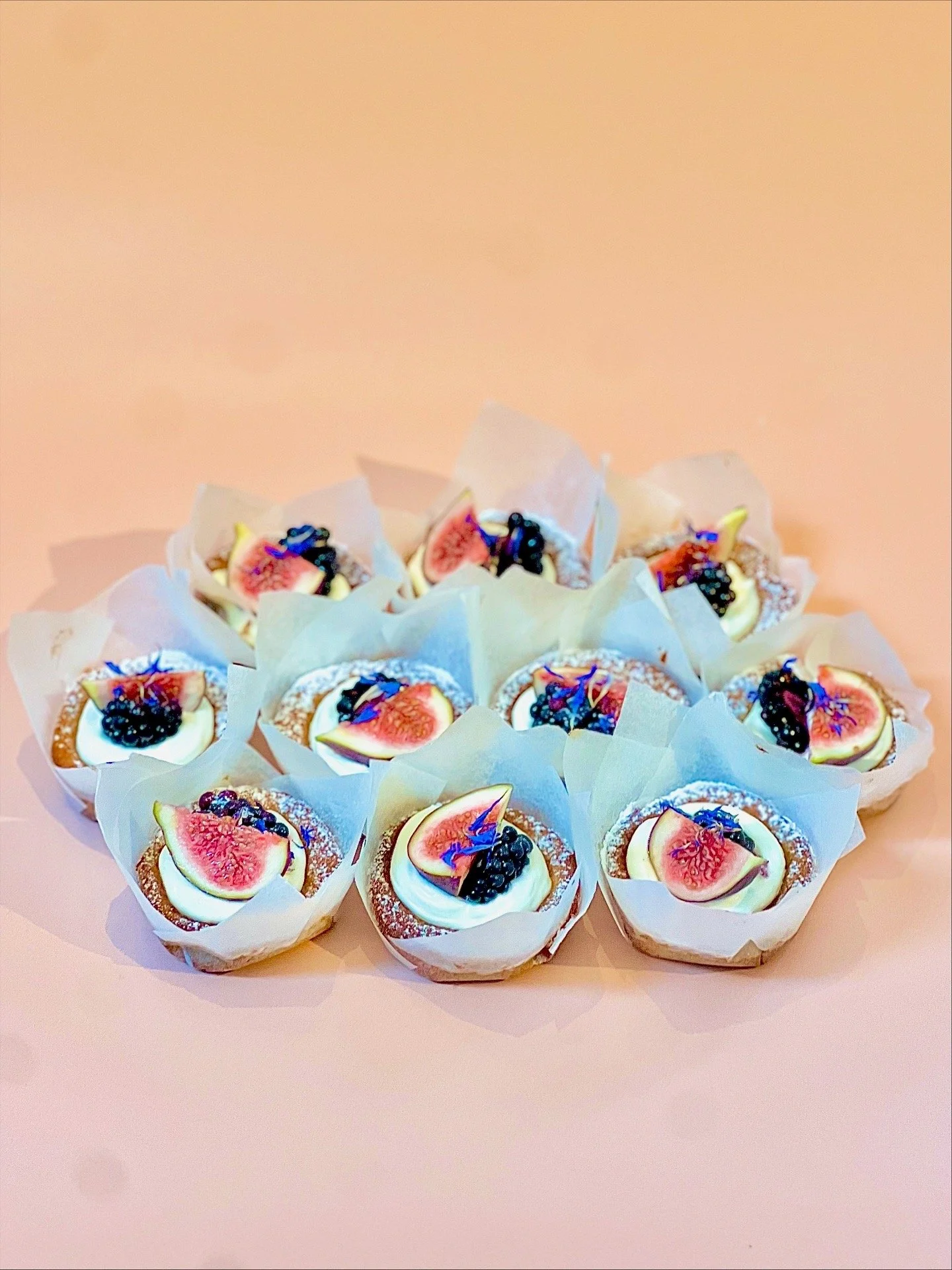 A collection of mini fruit tarts topped with figs, blueberries, and edible flowers on a pink surface with a plain pink background.
