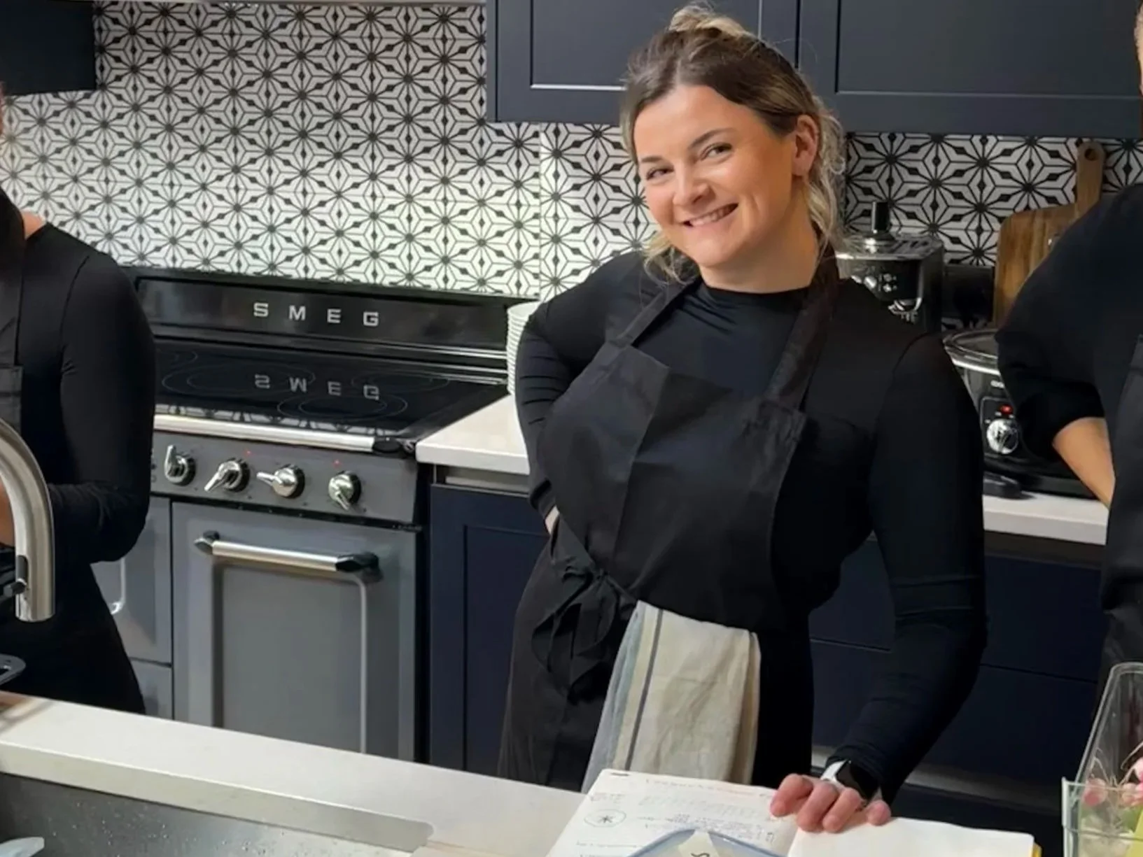A woman smiling in a kitchen, wearing a black apron, standing near a stove and countertop.