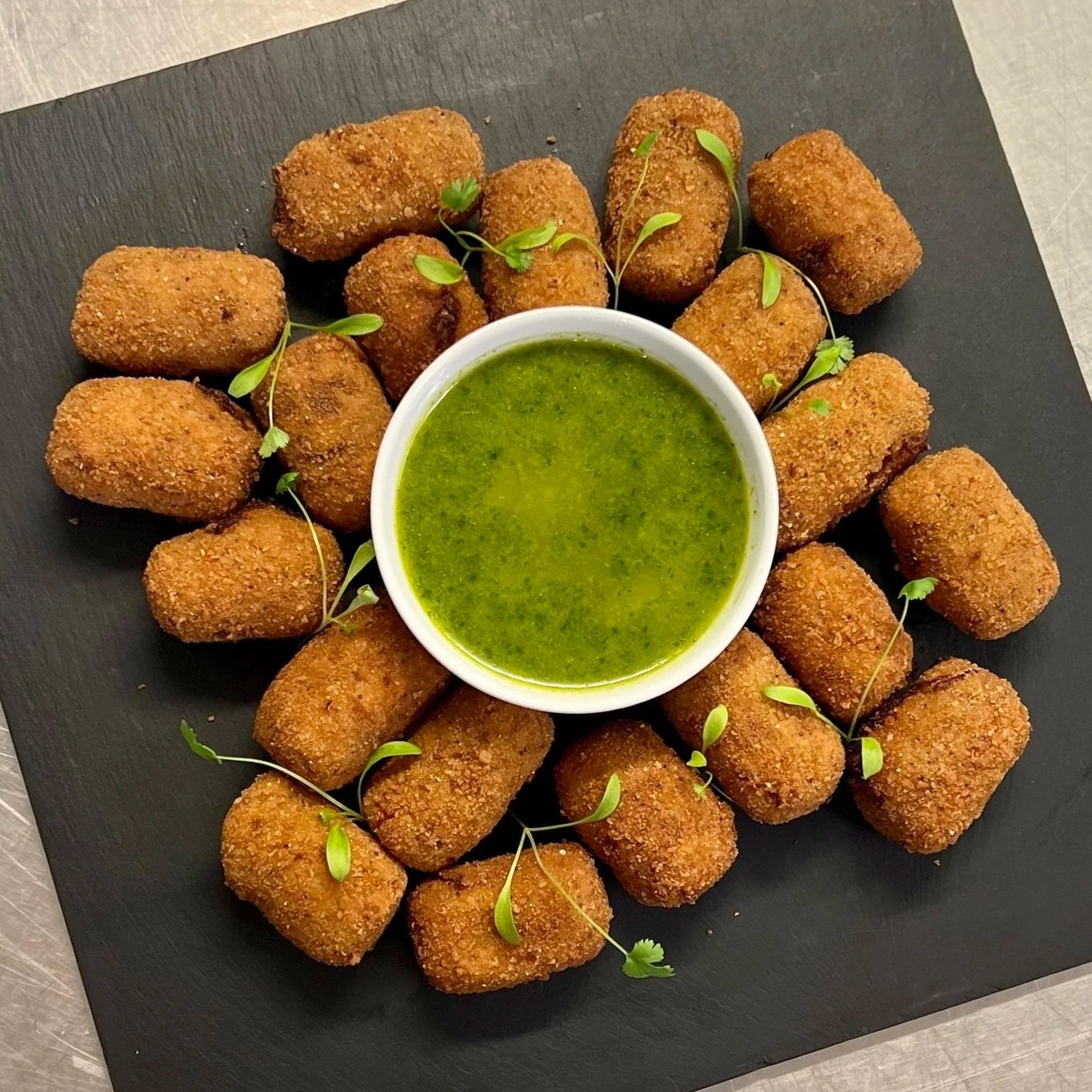 Fried croquettes arranged on a black tray with a small bowl of green dipping sauce in the center, garnished with microgreens.