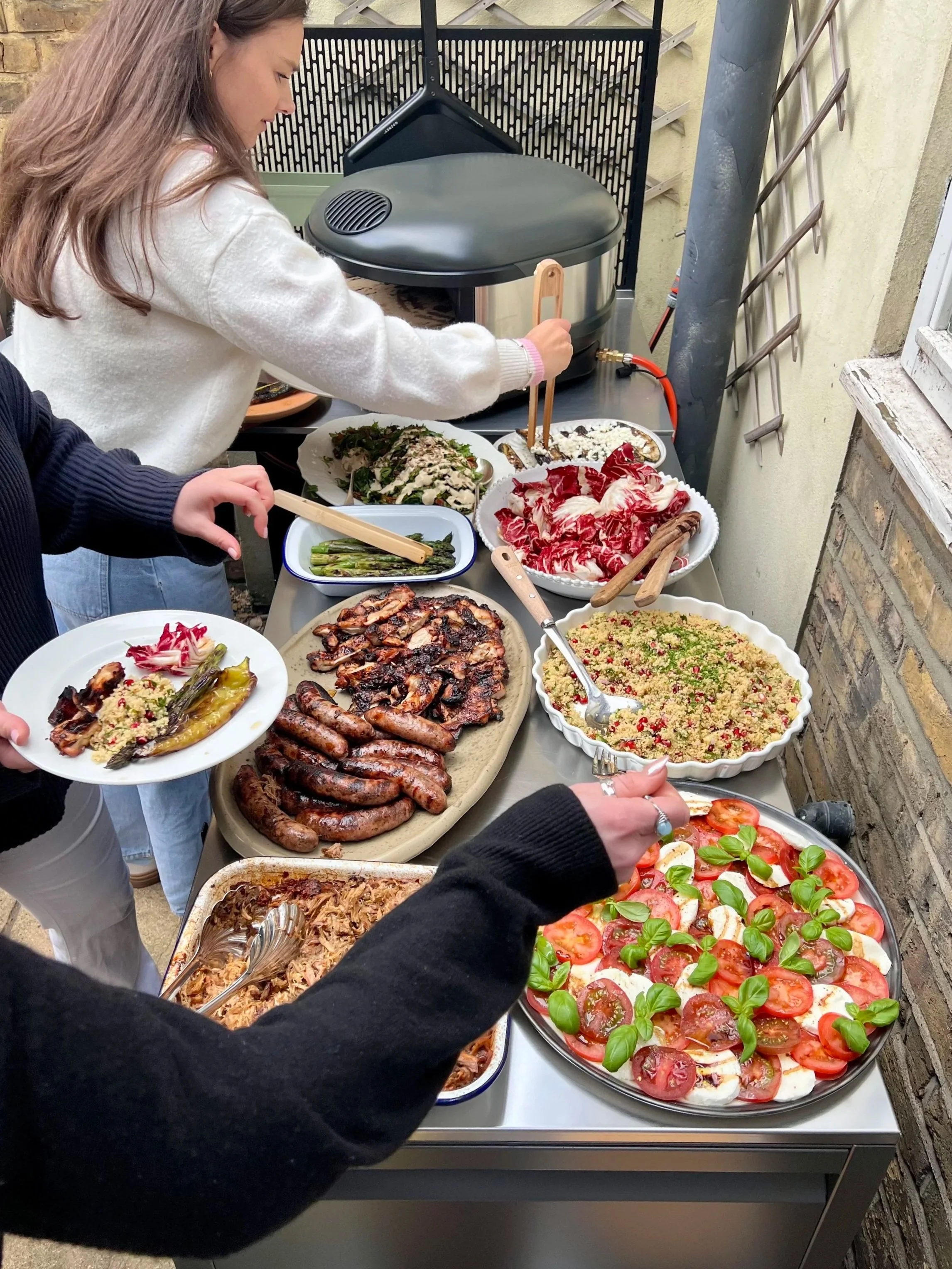 A person serving themselves food from an outdoor buffet table. The table has various dishes including a tomato and mozzarella salad, grilled sausages, sliced meats, pasta, and vegetables. Two other people are partially visible, one in a dark sweater 