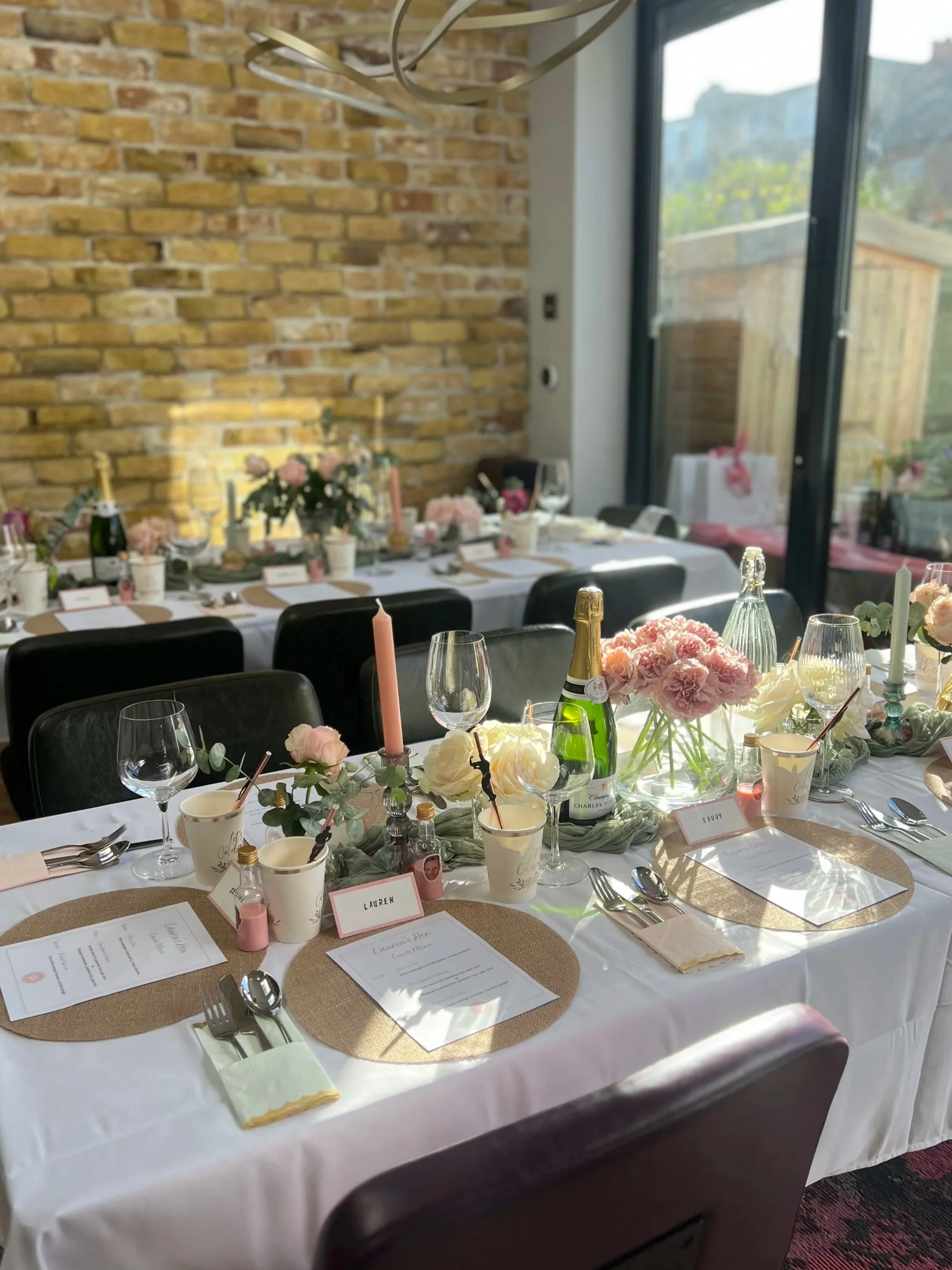 A decorated dining table set for a celebration, with floral arrangements, candles, wine bottles, and place cards, in a room with a brick wall and large windows.