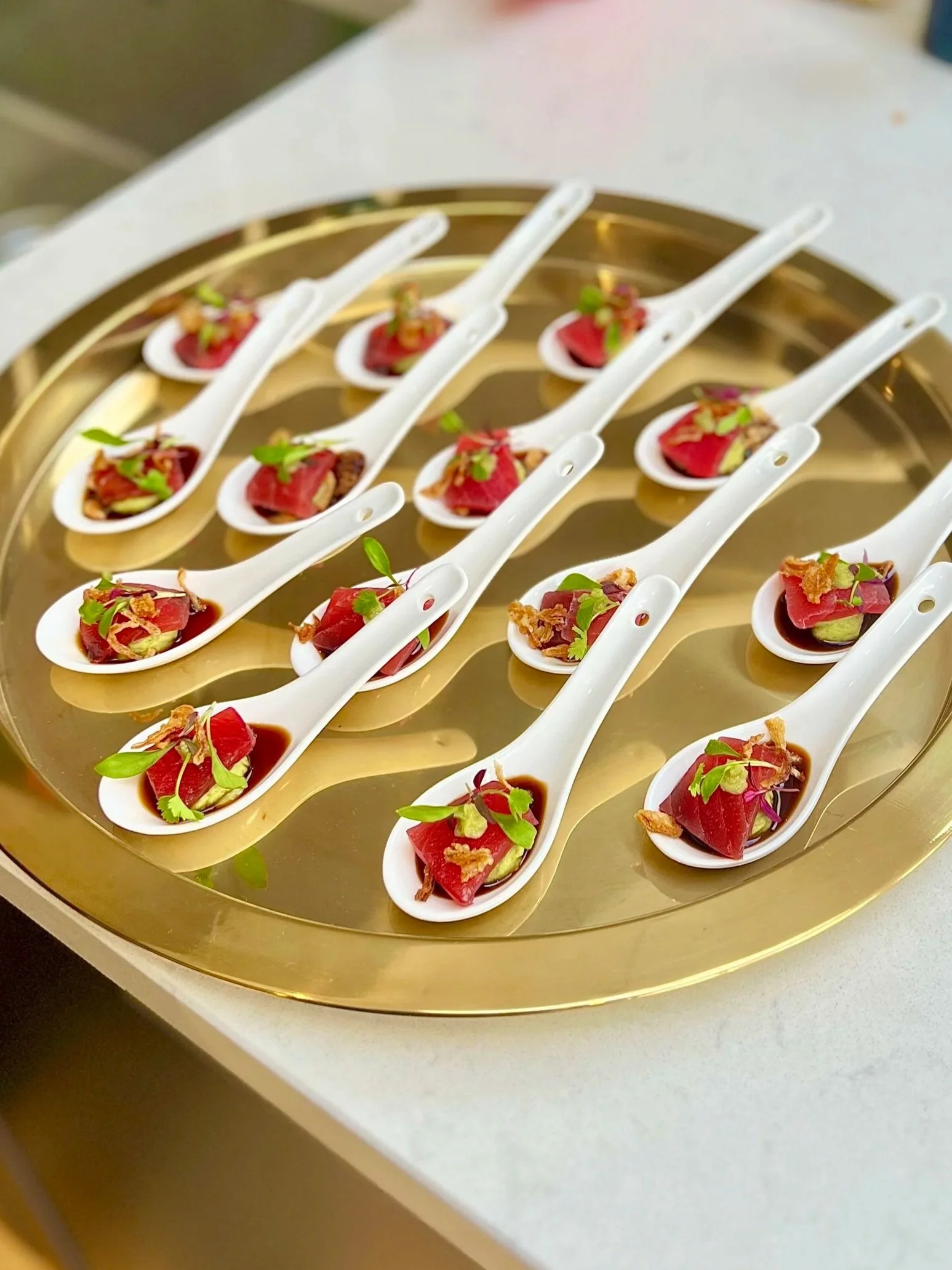 Small white spoons with a portion of raw tuna, garnished with microgreens, sitting on a gold-colored tray.