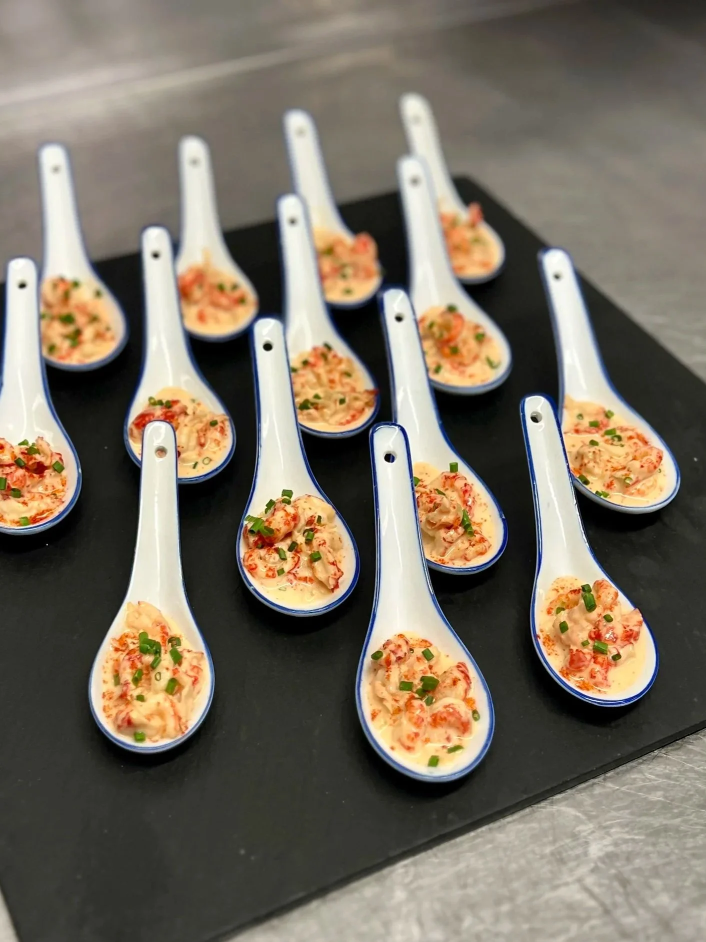Multiple small white ceramic spoons filled with a creamy seafood dish, garnished with chopped green onions, arranged on a black serving tray.