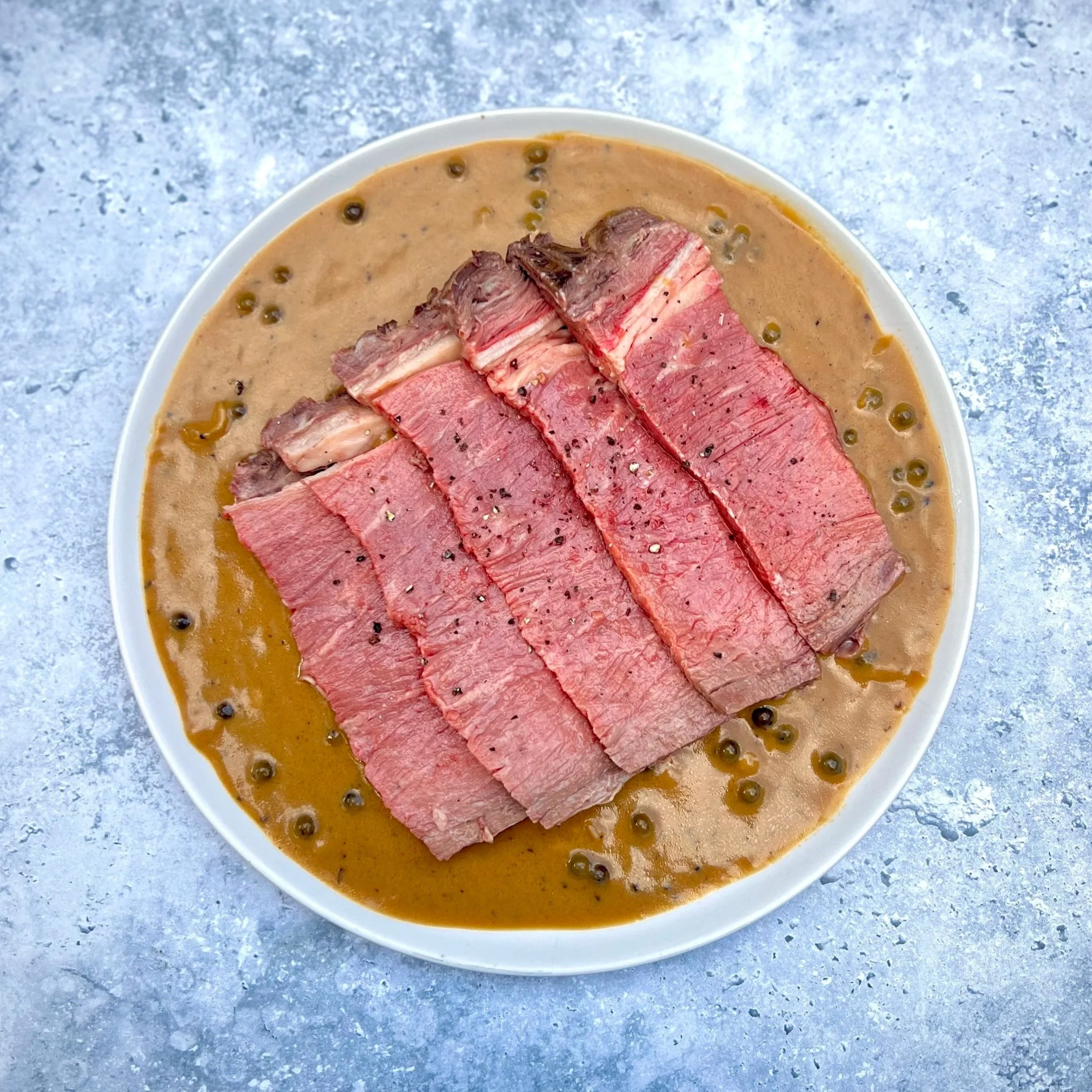 Sliced rare beef served on a white oval plate with green peppercorns in a beige sauce, placed on a gray marbled surface.