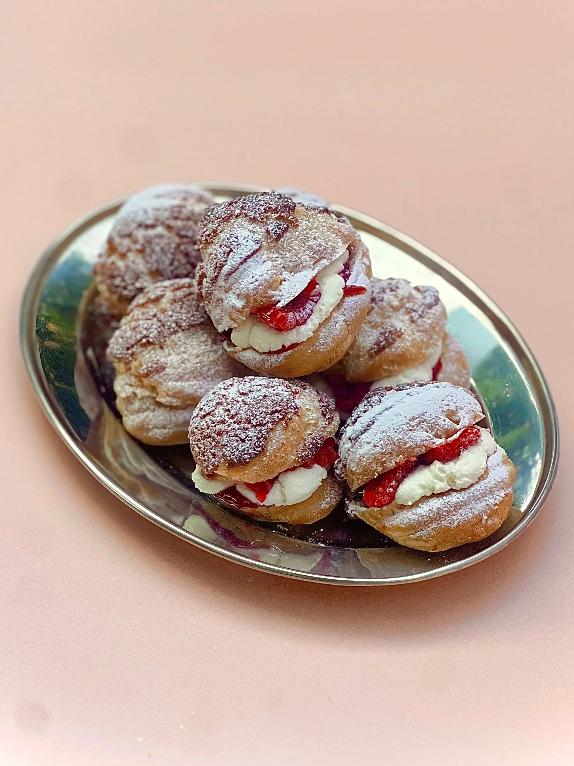 A silver tray holding several cream-filled eclairs topped with powdered sugar and garnished with red fruit, arranged on a pink surface.