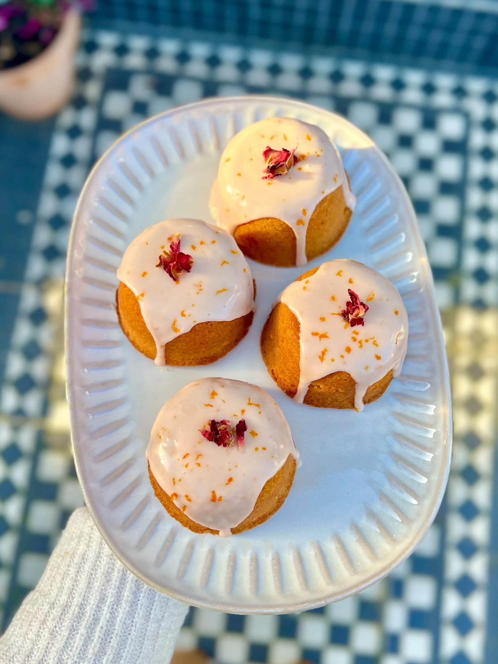 A white plate holding six small cupcakes with white icing, red flower petal decorations, and orange zest, held by a person wearing a white knit glove over a blue checkered surface.