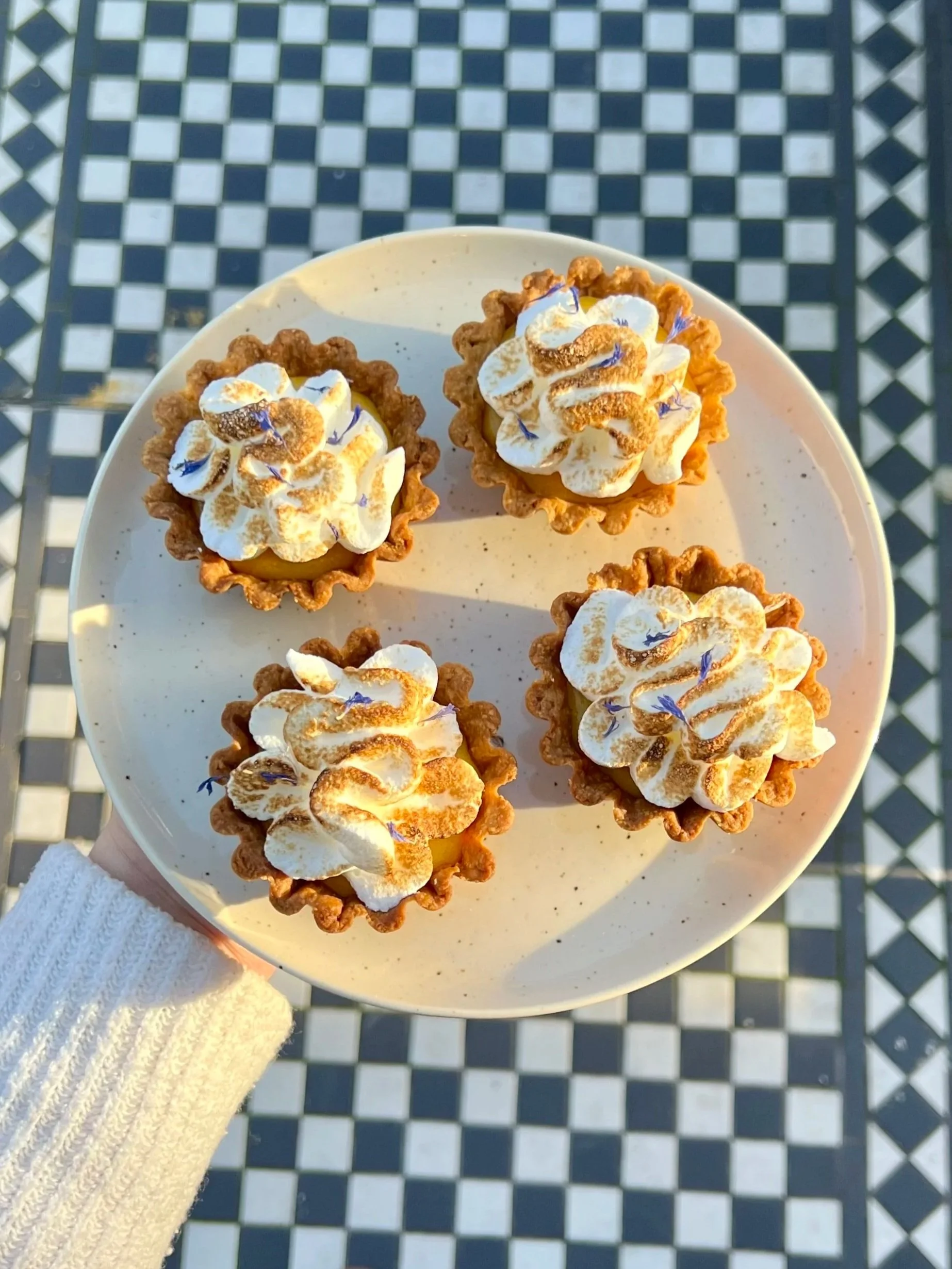 A white plate holding four lemon tarts topped with whipped cream and edible flowers, with a checkered black and white outdoor floor in the background.