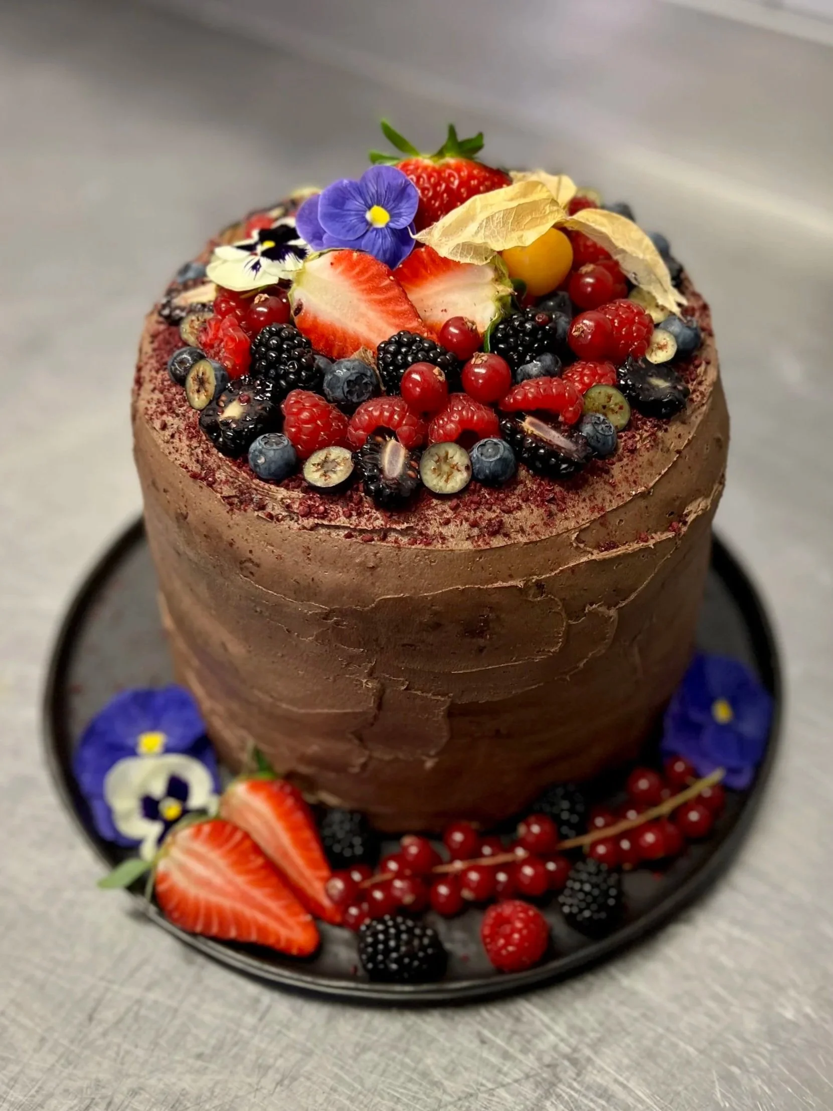 A cake decorated with fresh berries including strawberries, blueberries, raspberries, red currants, and blackberries, with edible flowers and dried leaves on top, placed on a black plate.
