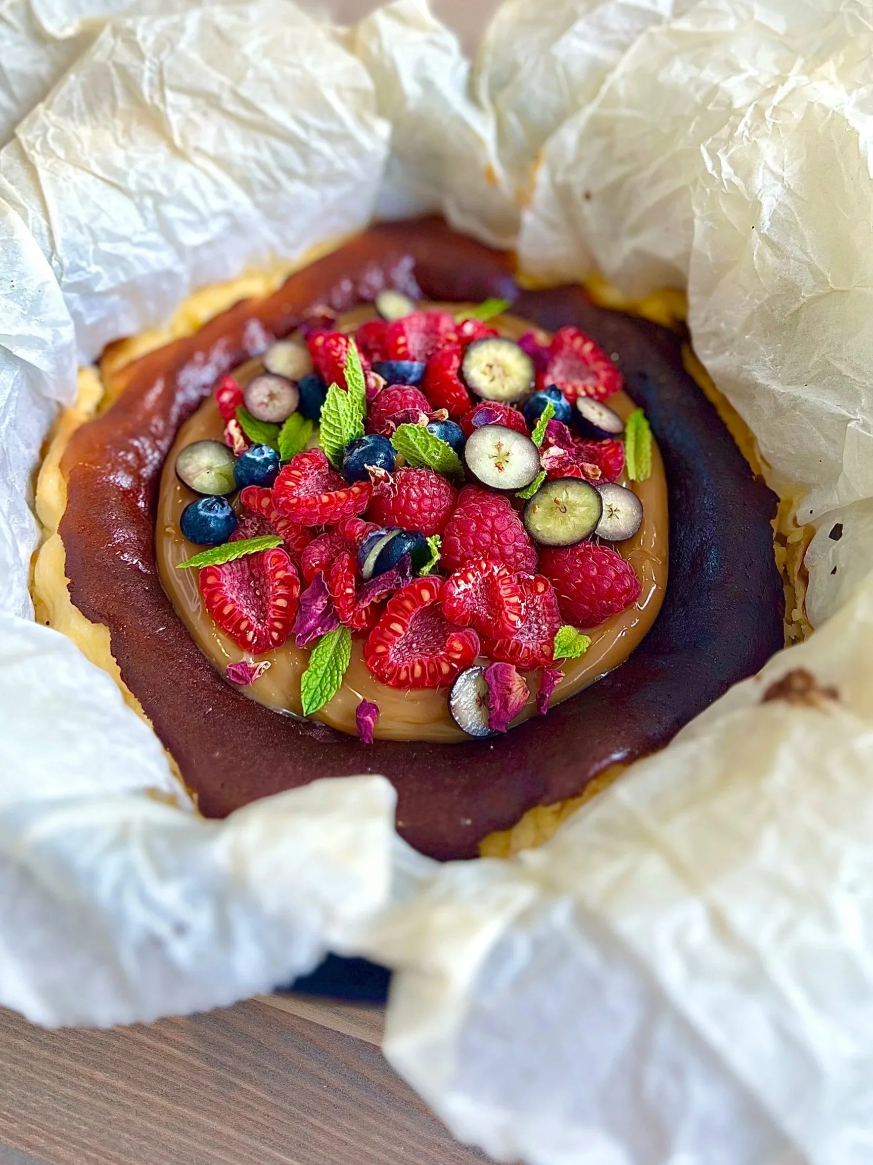 A baked good topped with mixed berries and fresh mint leaves, surrounded by crumpled parchment paper.