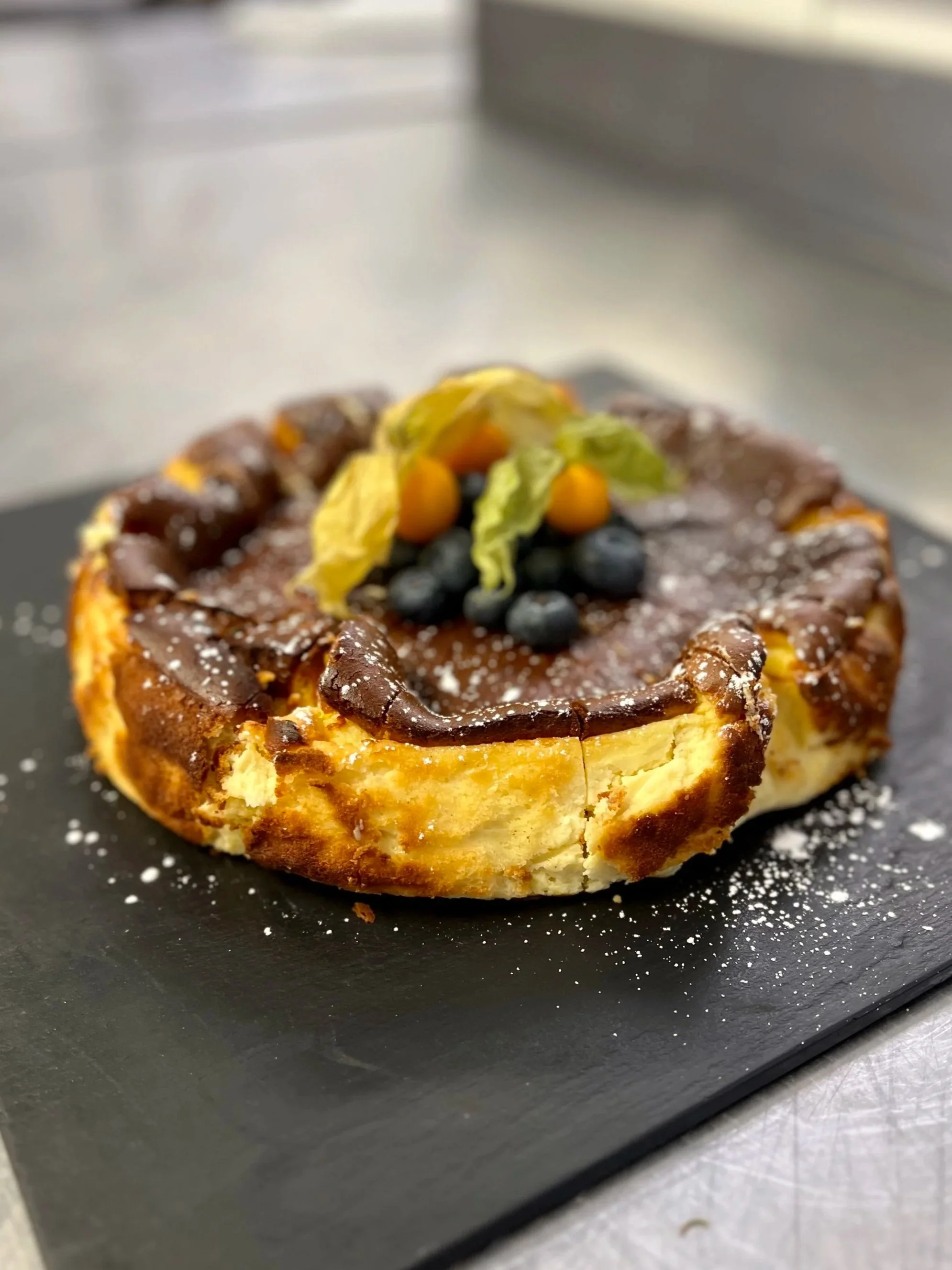 A round tart with a golden crust, topped with blueberries, physalis, and a drizzle of dark chocolate sauce, dusted with powdered sugar, on a black slate plate.