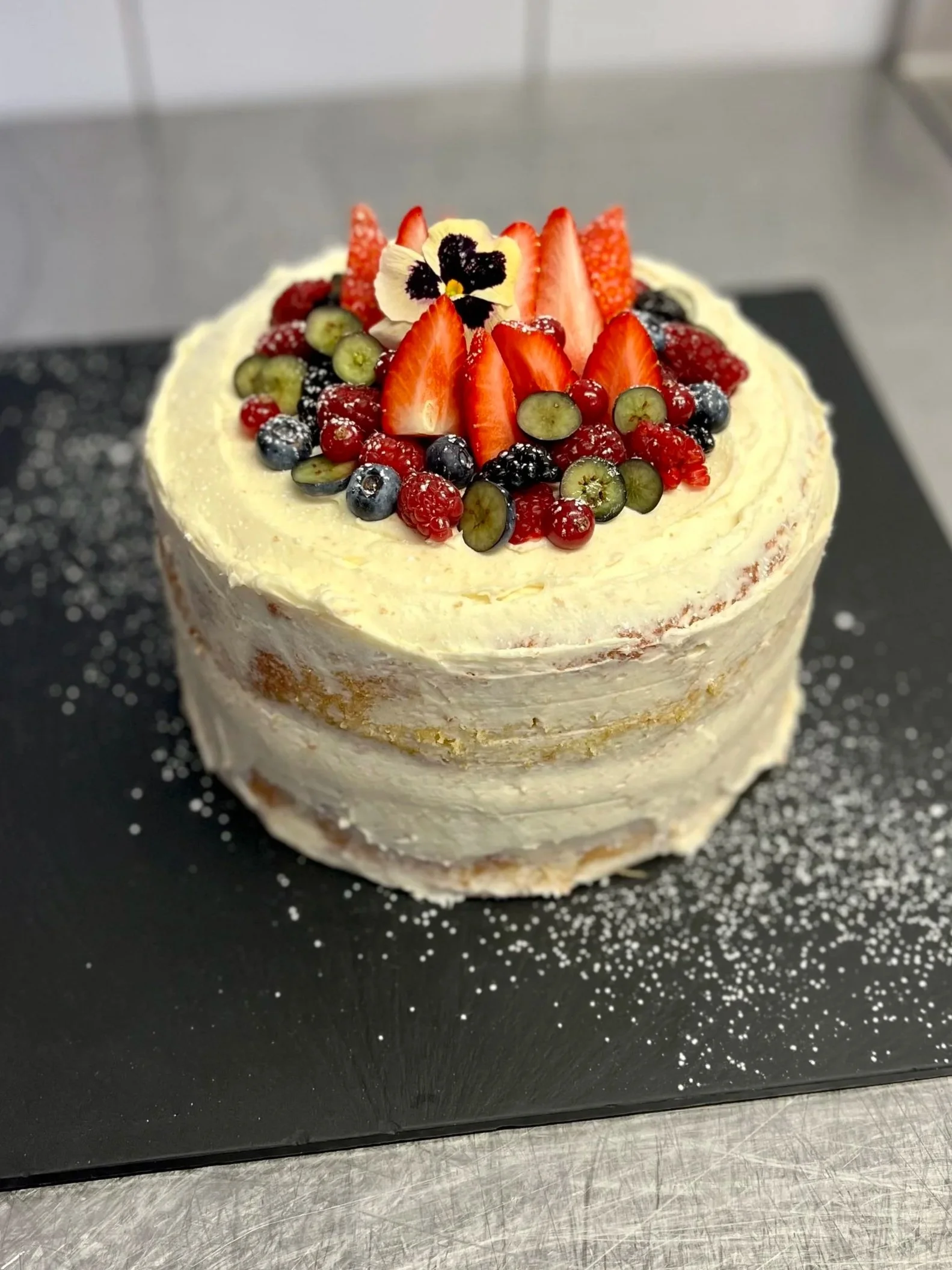 A layered cake decorated with various fresh berries and an edible flower on top, placed on a black surface sprinkled with powdered sugar.