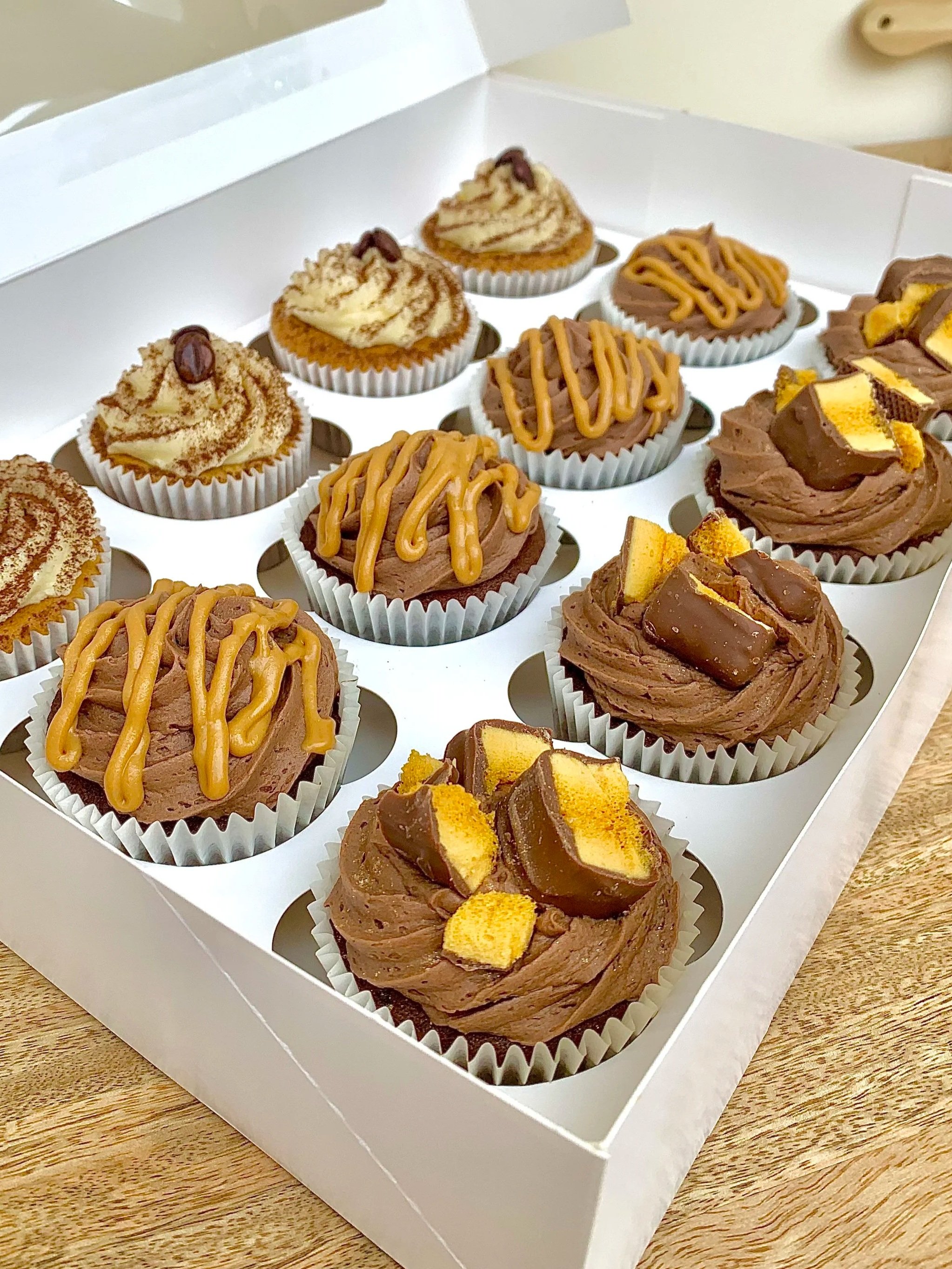 A white box with compartments holding assorted cupcakes on a wooden surface.