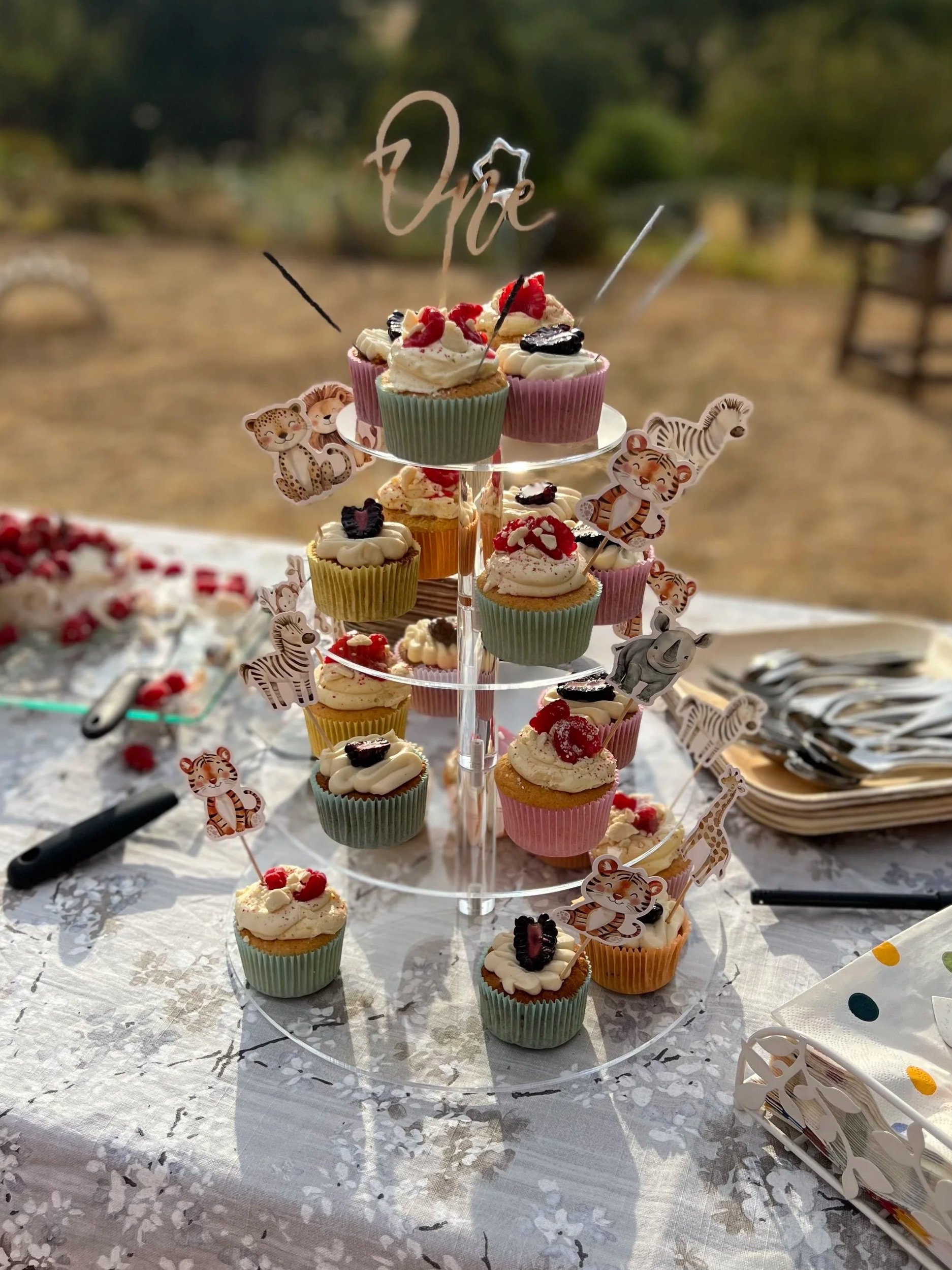Tiered stand filled with decorated cupcakes topped with whipped cream, berries, and animal-themed toppers, set on a table during an outdoor event.