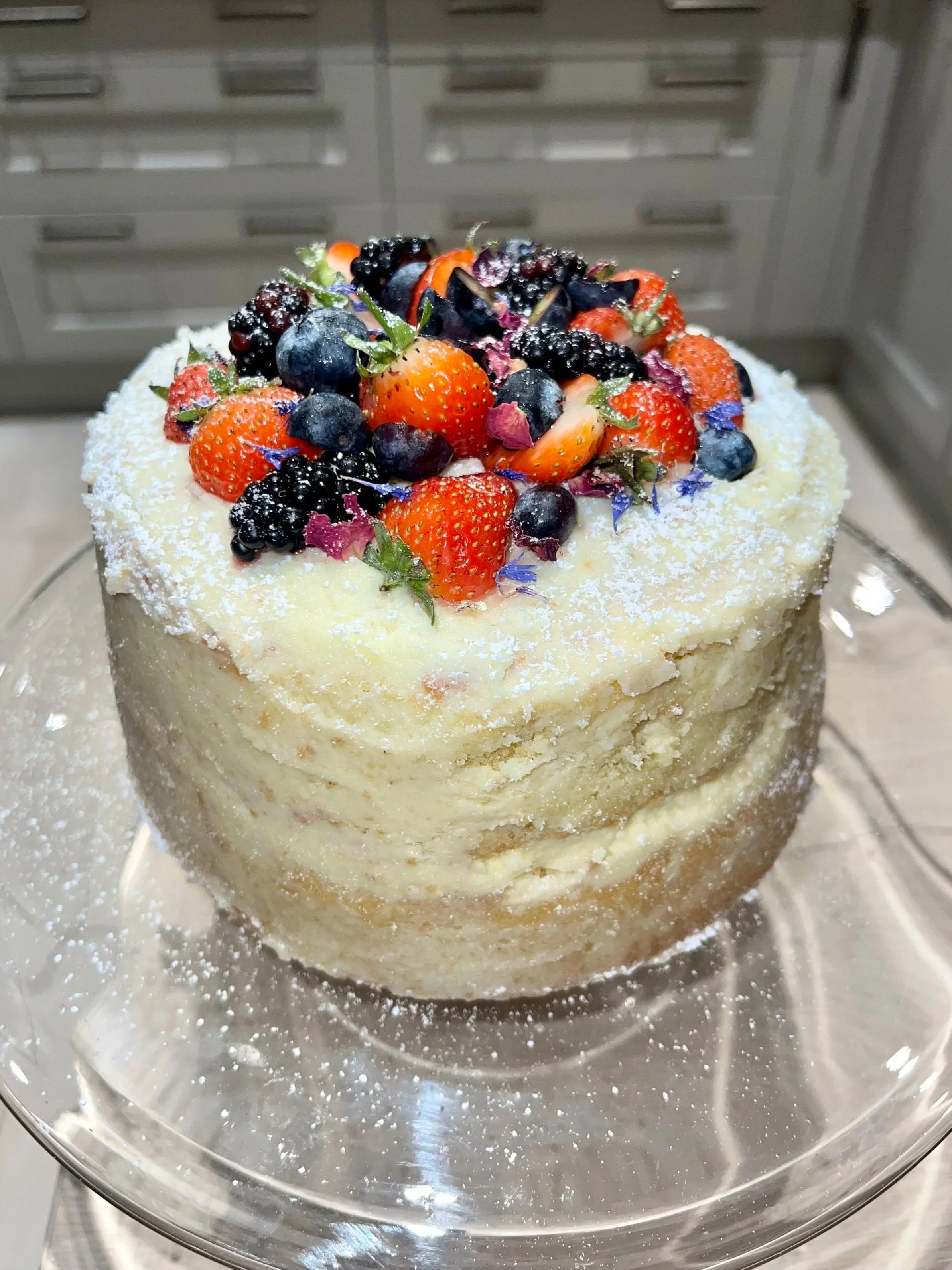 A cake topped with fresh strawberries, blueberries, blackberries, and edible flowers, with powdered sugar dusted on top, inside a glass cake cover.