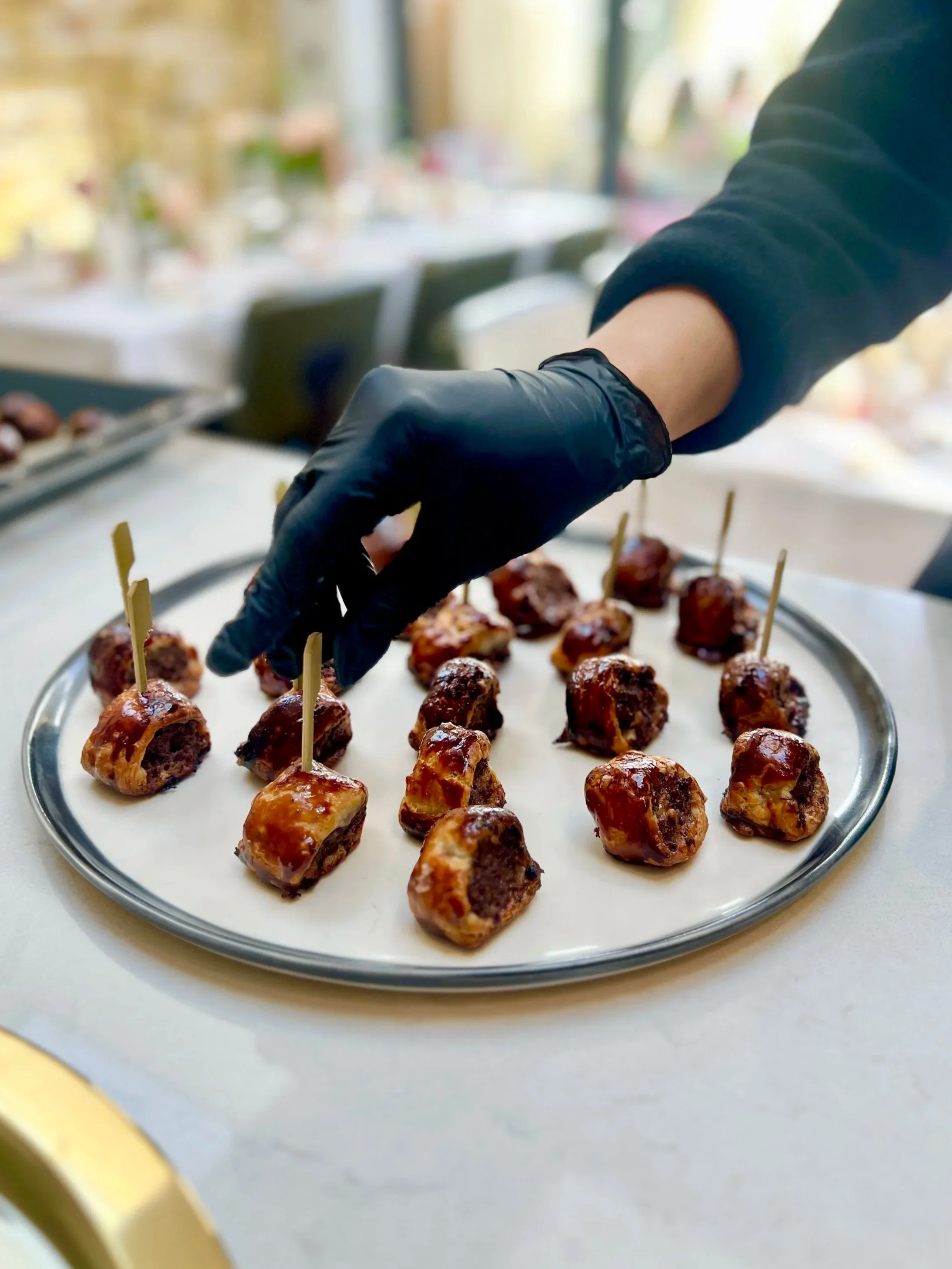 Hand wearing a black glove serving glazed meatballs on a silver tray.