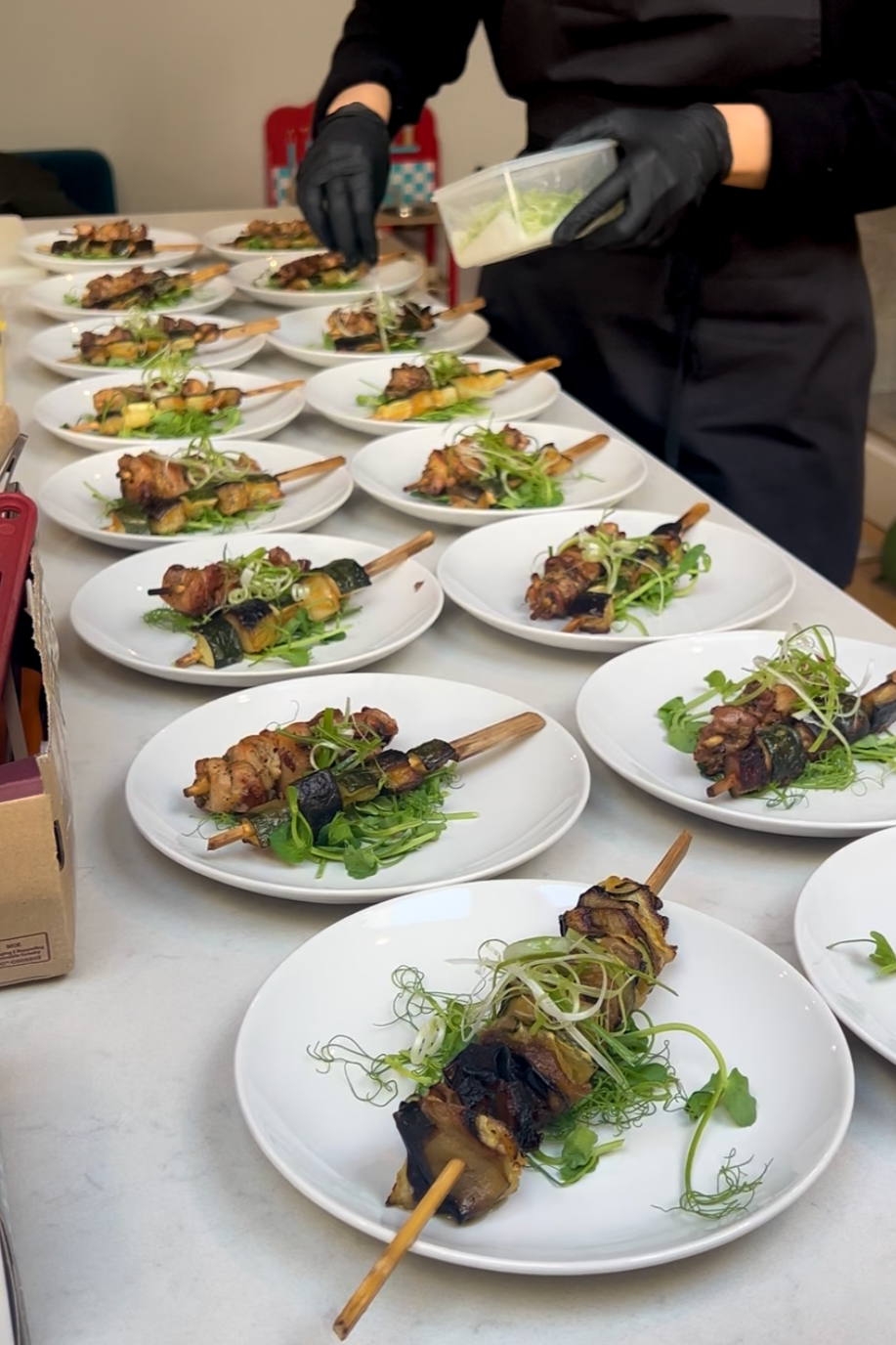 Multiple plates of grilled meat and vegetables on a table, with a chef in black gloves preparing the dishes.
