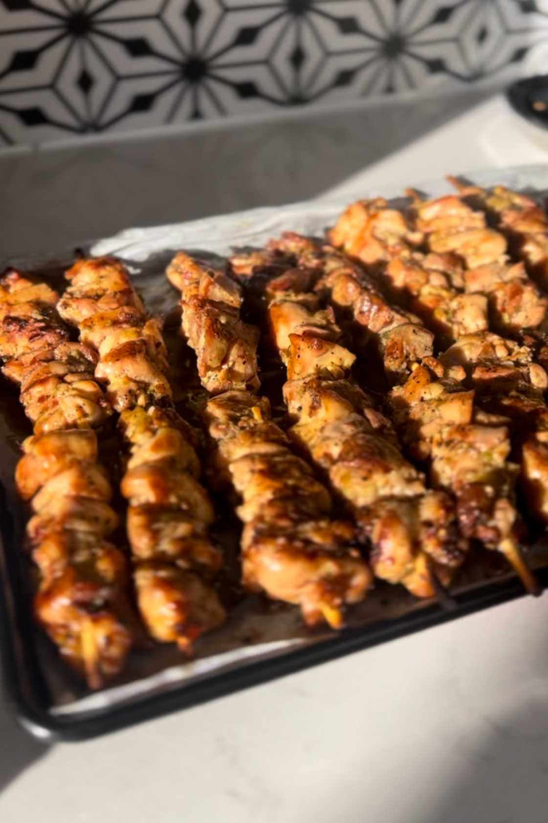 Close-up of grilled chicken skewers on a baking sheet lined with parchment paper, with a patterned background.