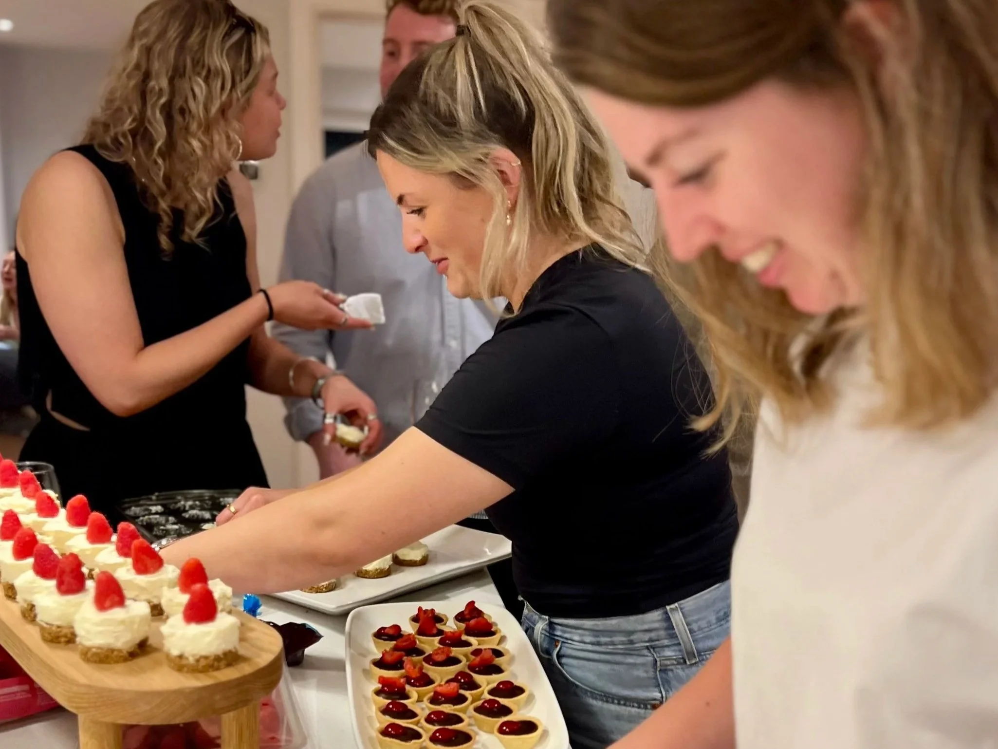 People serving and enjoying desserts at a gathering, including a cake topped with strawberries and cupcakes with strawberries and berry toppings.