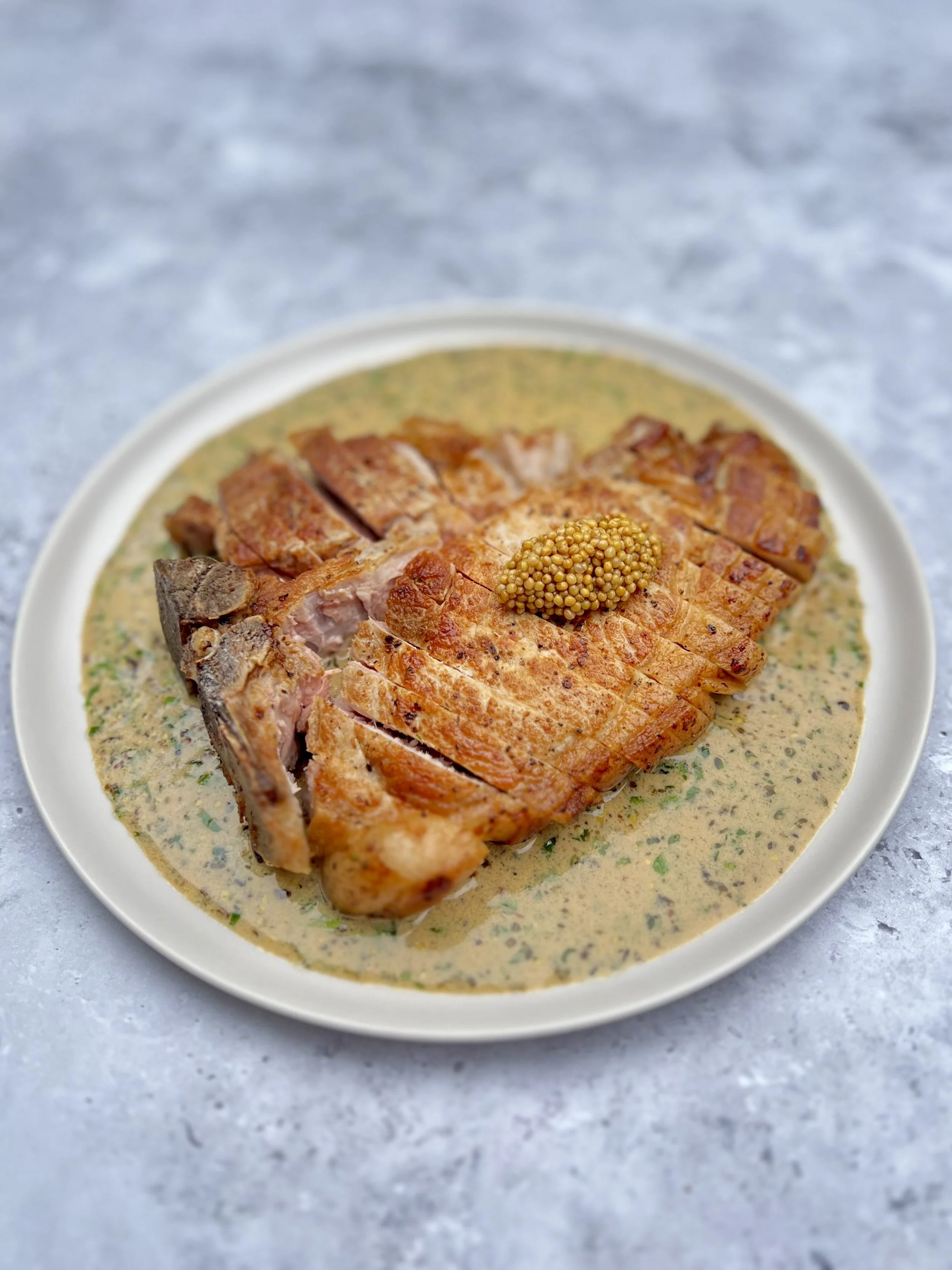 A piece of cooked salmon fillet with crispy skin on a white bowl filled with creamy herb sauce, garnished with a small dollop of mustard seeds, on a light gray surface.