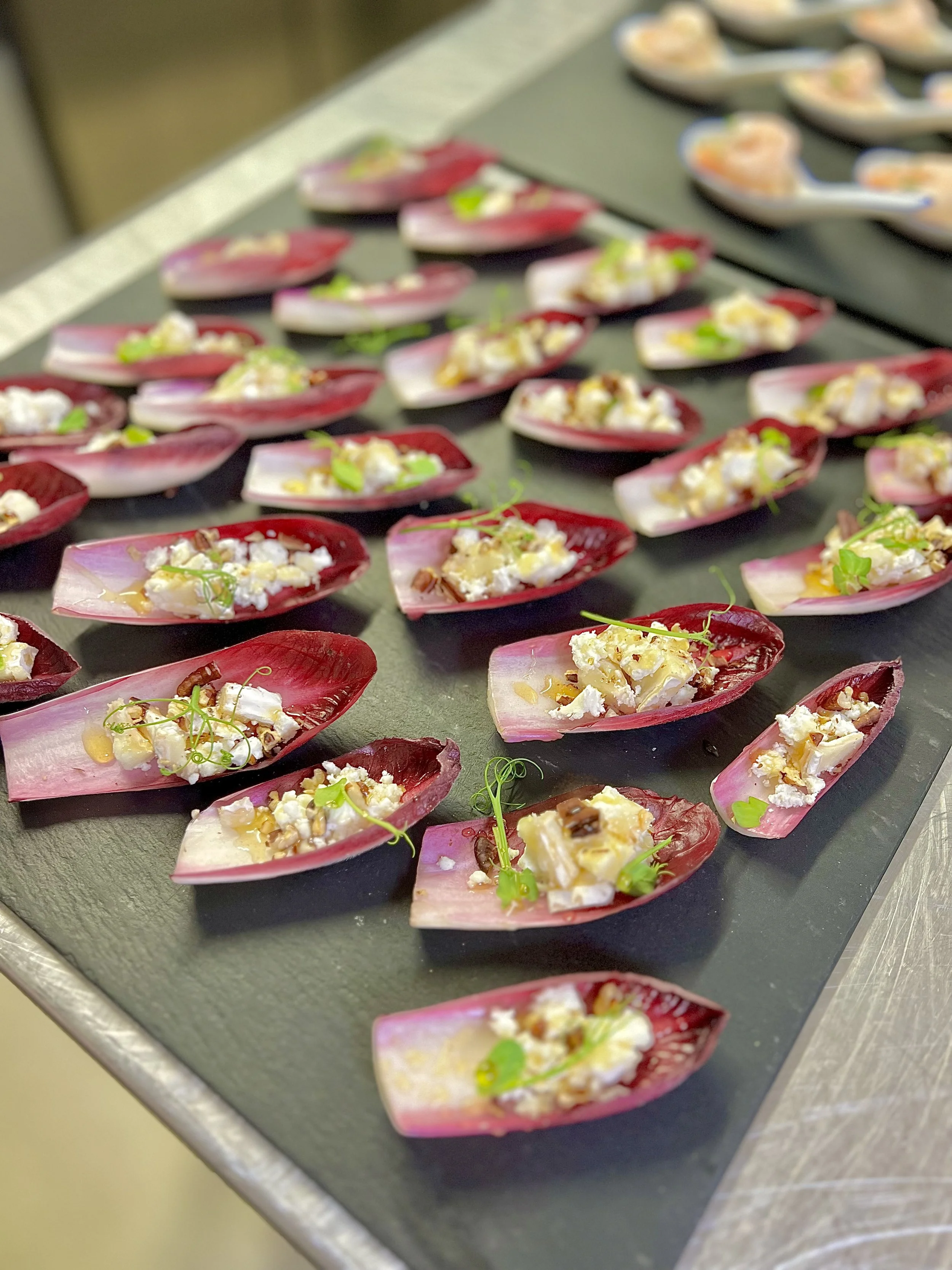 Close-up of bite-sized appetizer trays with pink radicchio chips filled with cheese, herbs, and garnished with microgreens, on a black serving tray.