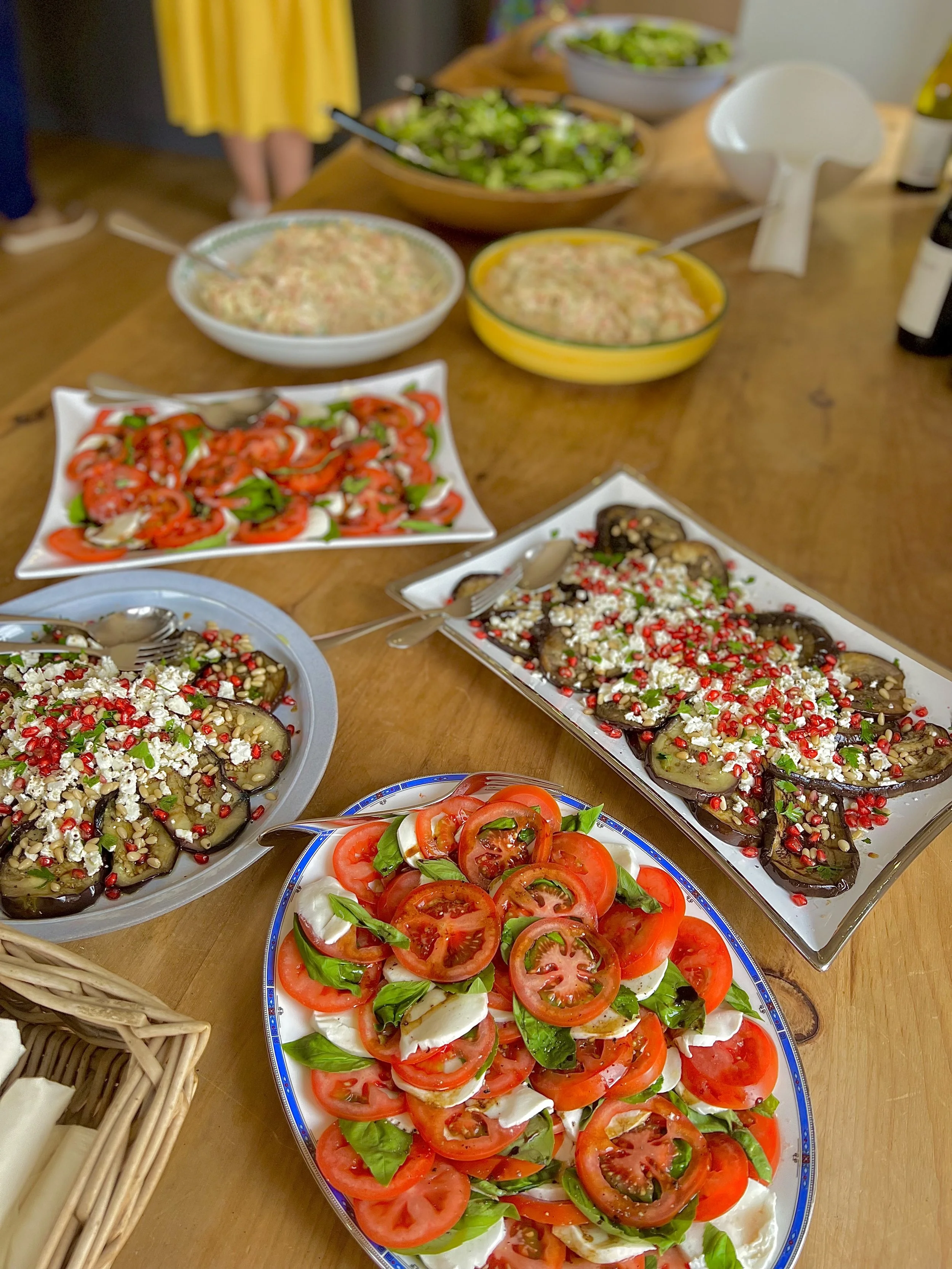 A table set with various dishes, including a Caprese salad with sliced tomatoes, mozzarella, and basil, eggplant topped with pomegranate seeds, a dish with a creamy mixture, and a salad with mixed greens, along with utensils and bowls, in a casual di