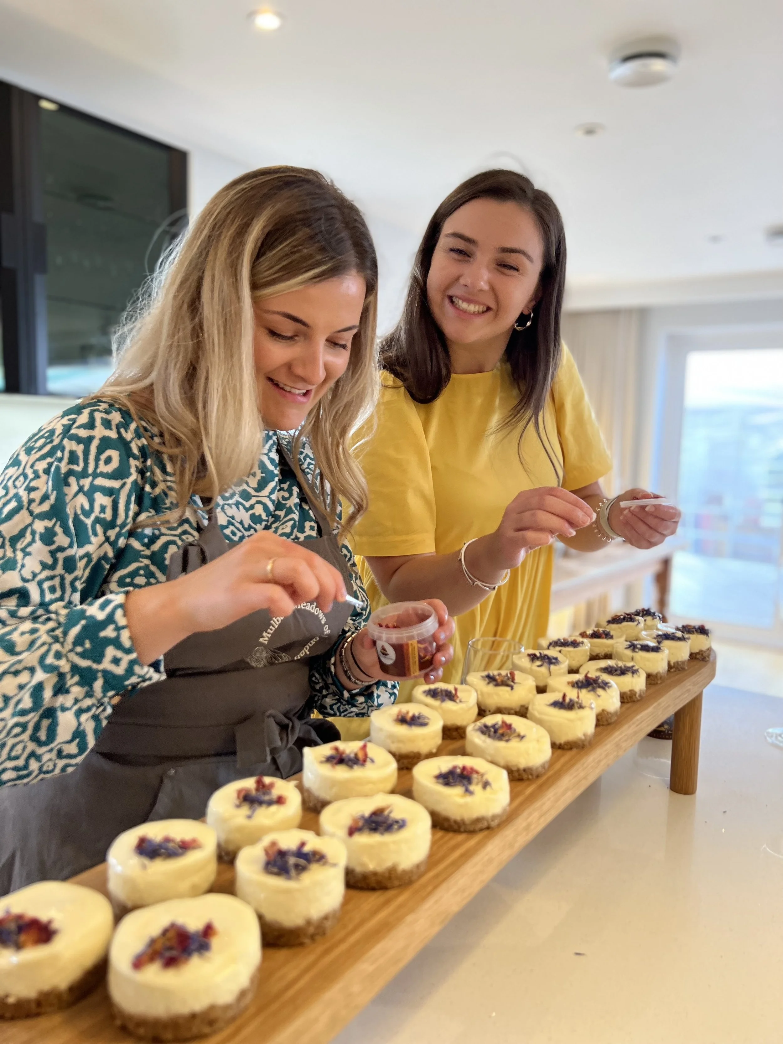 Two women decorating mini cheesecakes on a wooden tray in a bright indoor setting.