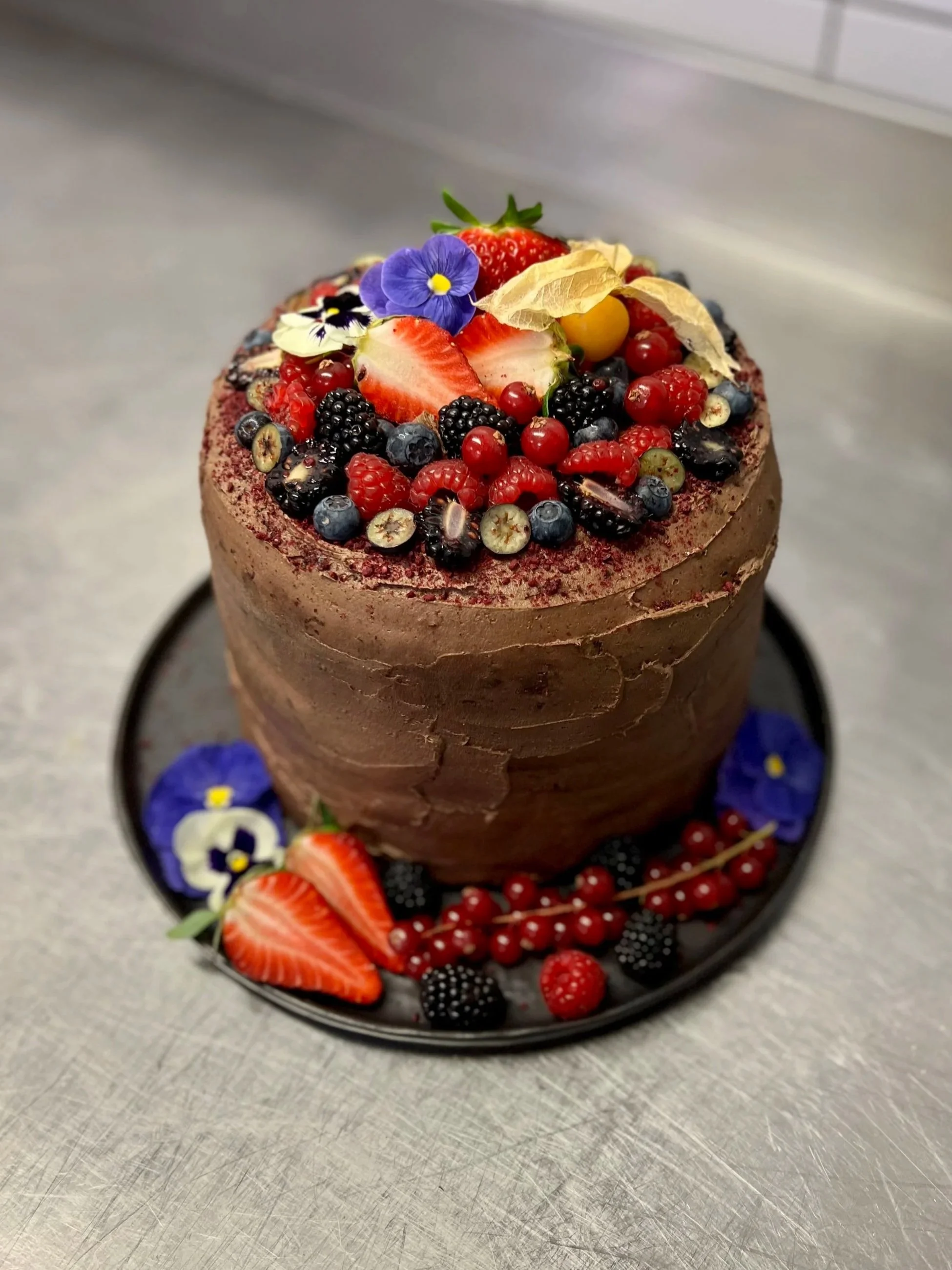 Chocolate cake topped with strawberries, blueberries, blackberries, red currants, and edible flowers, displayed on a black cake stand.
