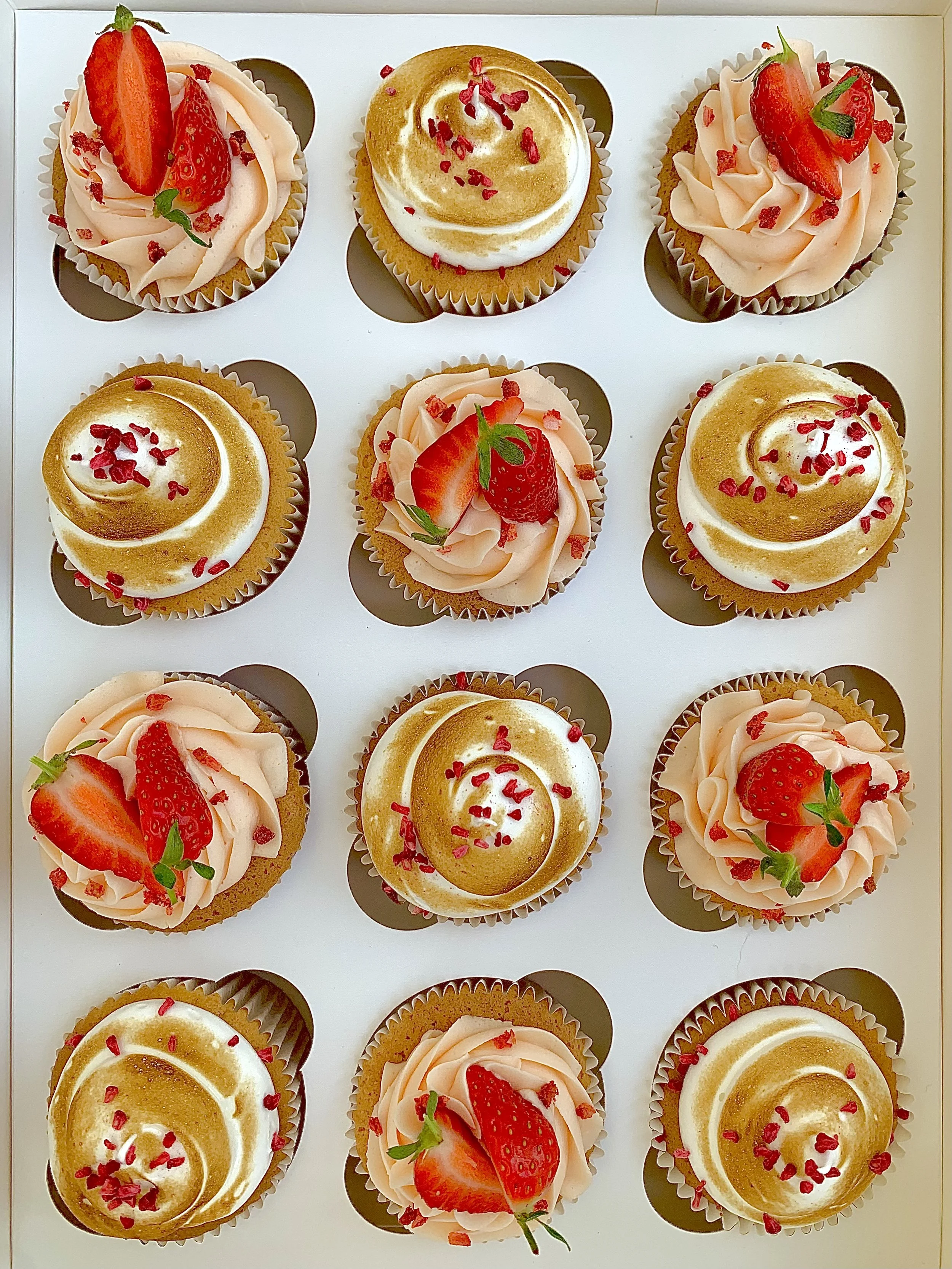 A box of twelve strawberry cupcakes topped with pink frosting, fresh strawberries, and red sprinkles.
