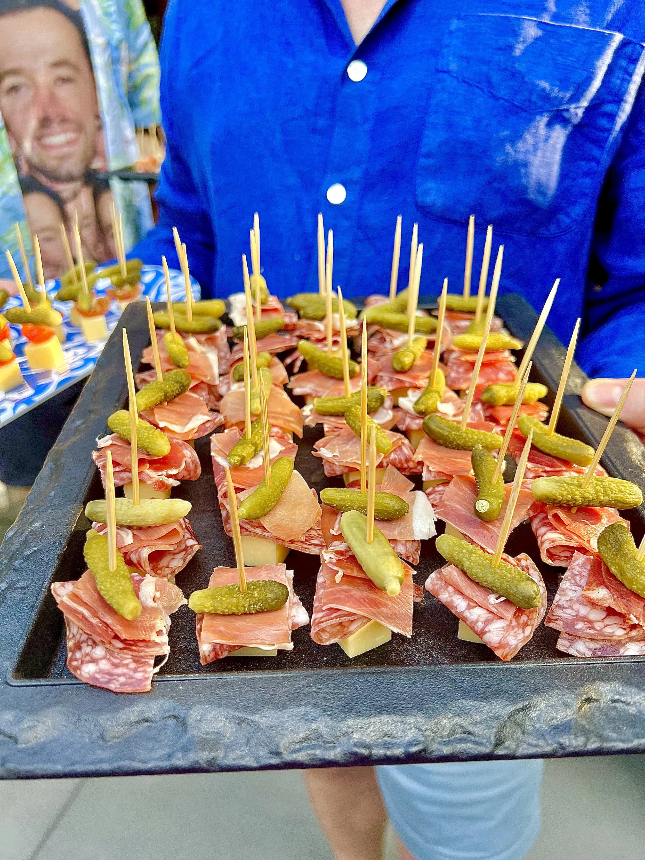 Tray of small appetizers with slices of meat, cheese, and pickles skewered with toothpicks, on a black serving tray.