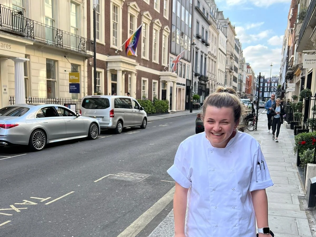 A young female chef in a white uniform laughing on a city street with parked cars and buildings in the background.
