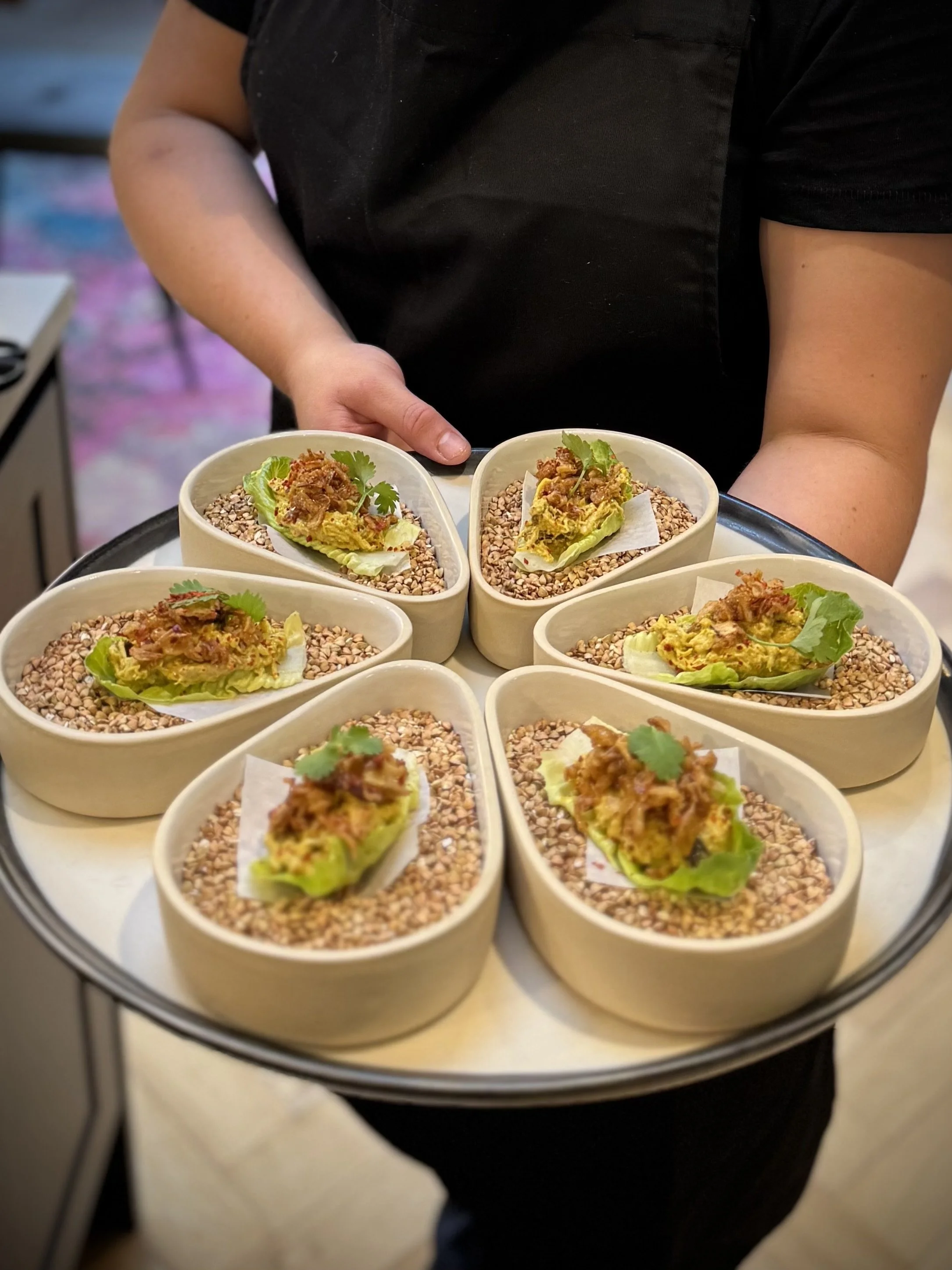 A person in a black shirt holding a tray with multiple small bowls of food topped with greens.