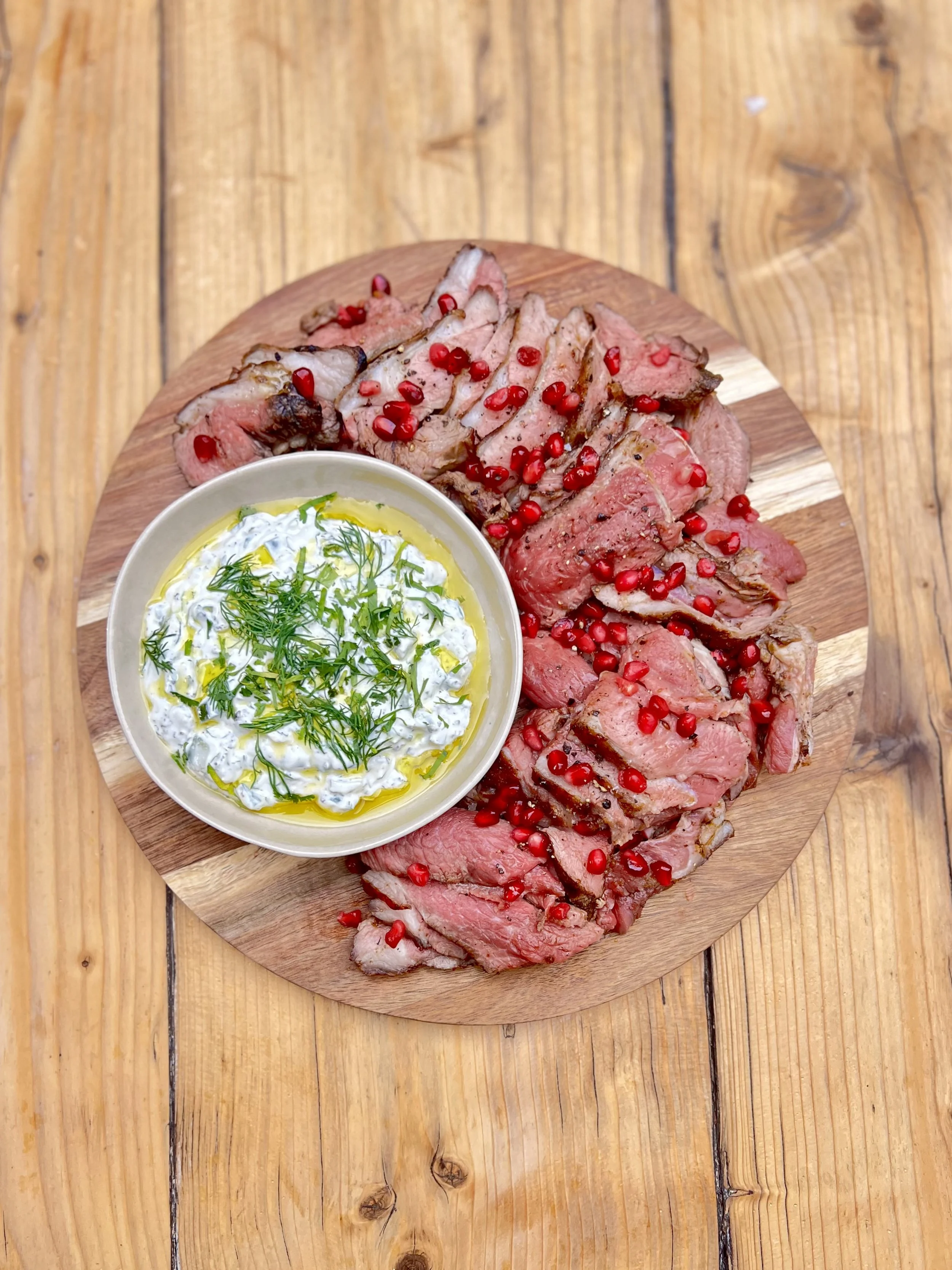 Sliced grilled steak with pomegranate seeds served on a wooden platter with a side of creamy dill sauce