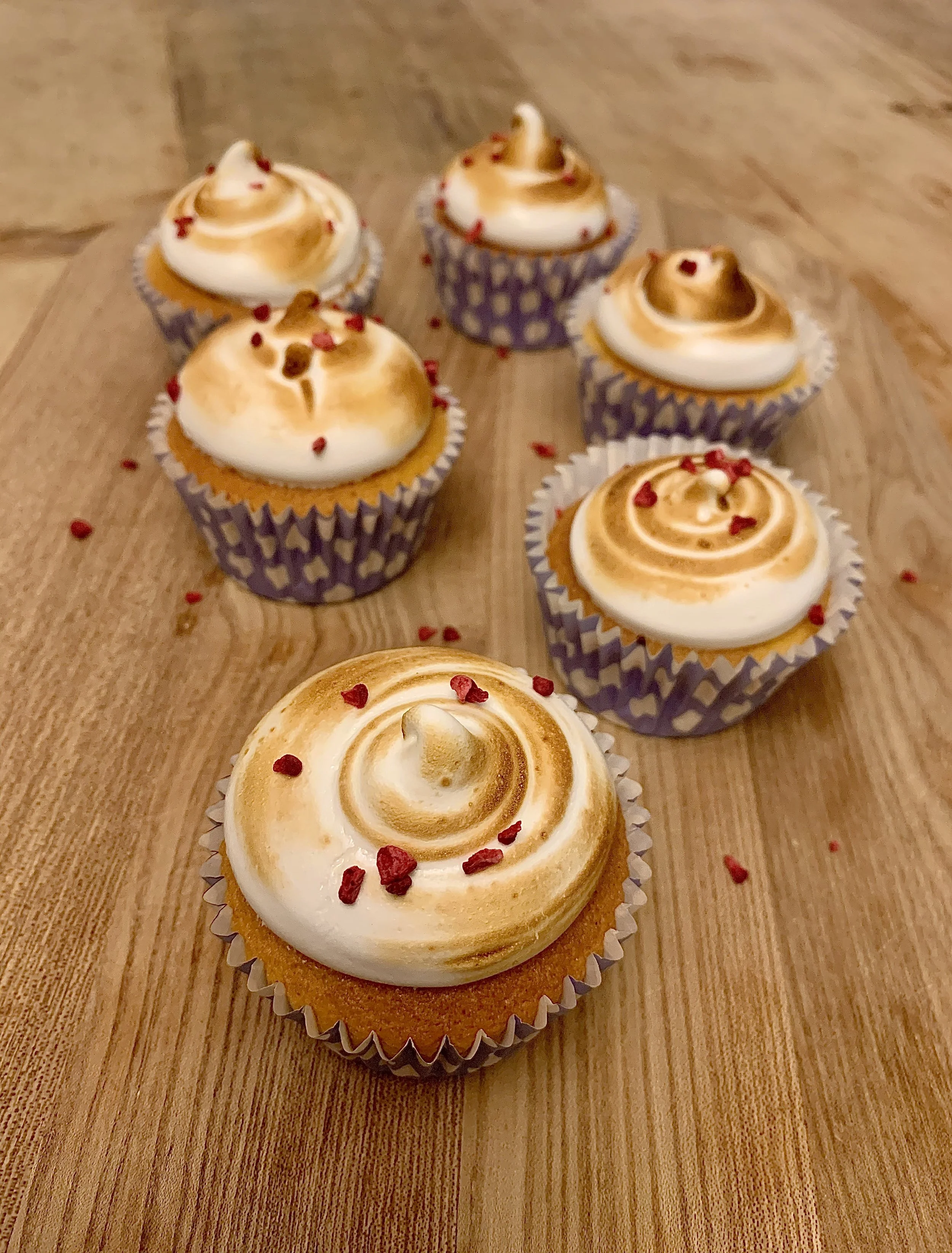 Five cupcakes with white and light brown swirled frosting and red sprinkles on a wooden surface.