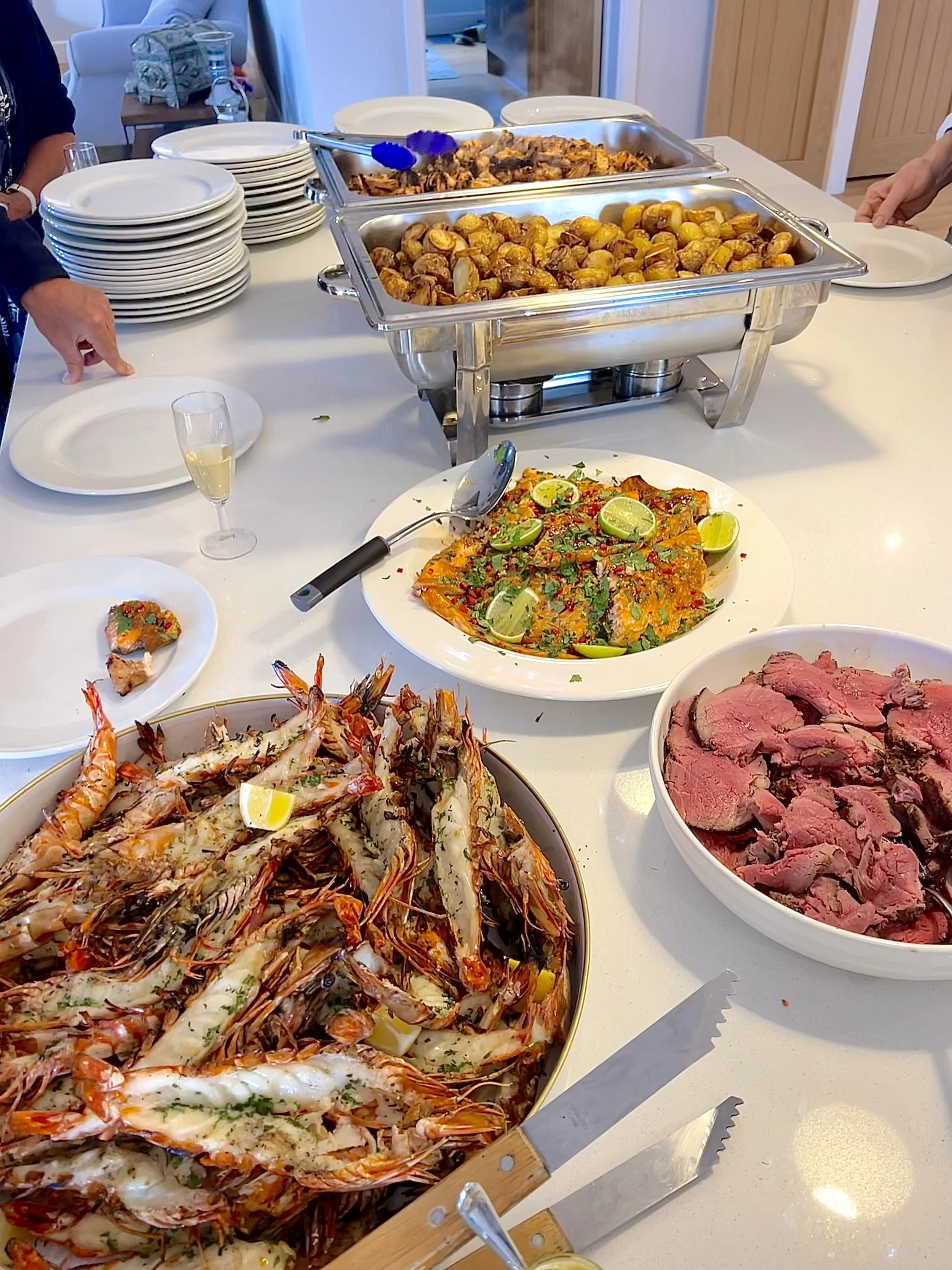 Buffet table with various dishes including a large plate of cooked shrimp, bowls of sliced roast beef, a grilled fish dish garnished with lime, roasted potatoes, and other foods. Plates and utensils are set nearby.