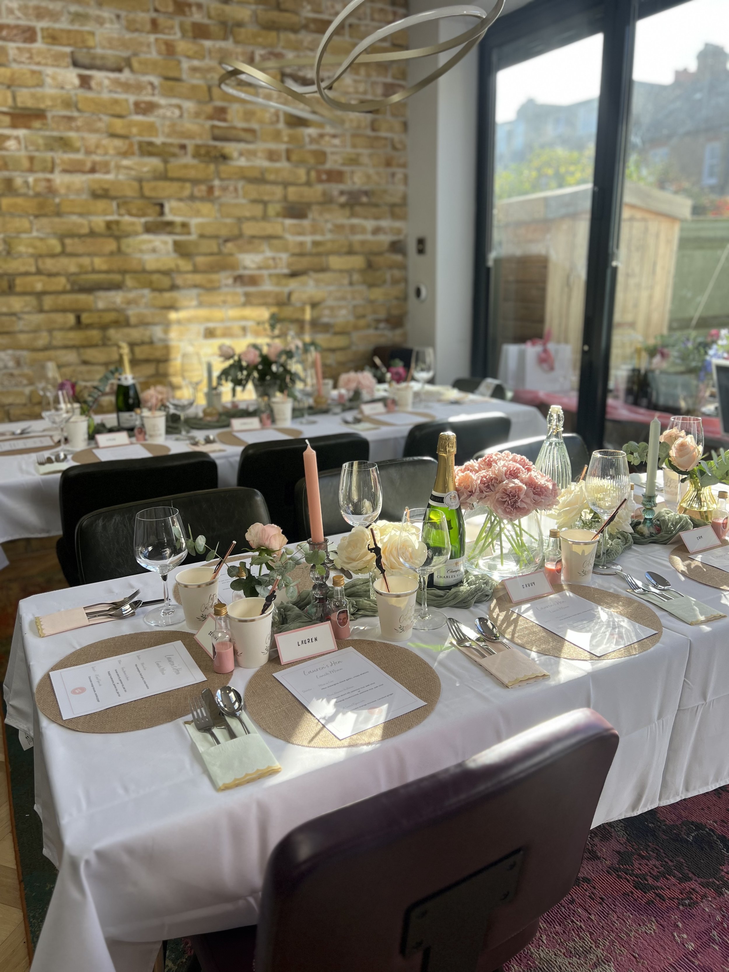 A decorated banquet table with pink, white, and green floral arrangements, wine bottles, glasses, candles, and place cards, set for a celebration or event in a room with a brick wall, large windows, and a chandelier.