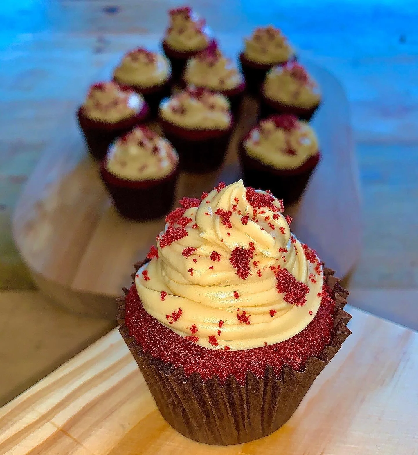 A cupcake with red velvet cake, cream cheese frosting, and red sprinkles in the foreground, and multiple cupcakes with frosting and sprinkles on a tray in the background.