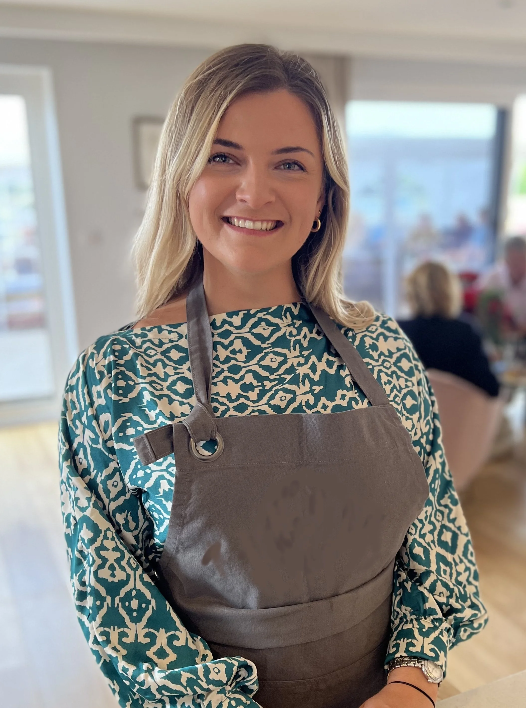 A smiling woman with blonde hair wearing a patterned blue and white top and a brown apron inside a kitchen or cafe setting.