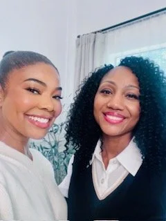 Two women smiling for a selfie indoors, standing near a window with white curtains