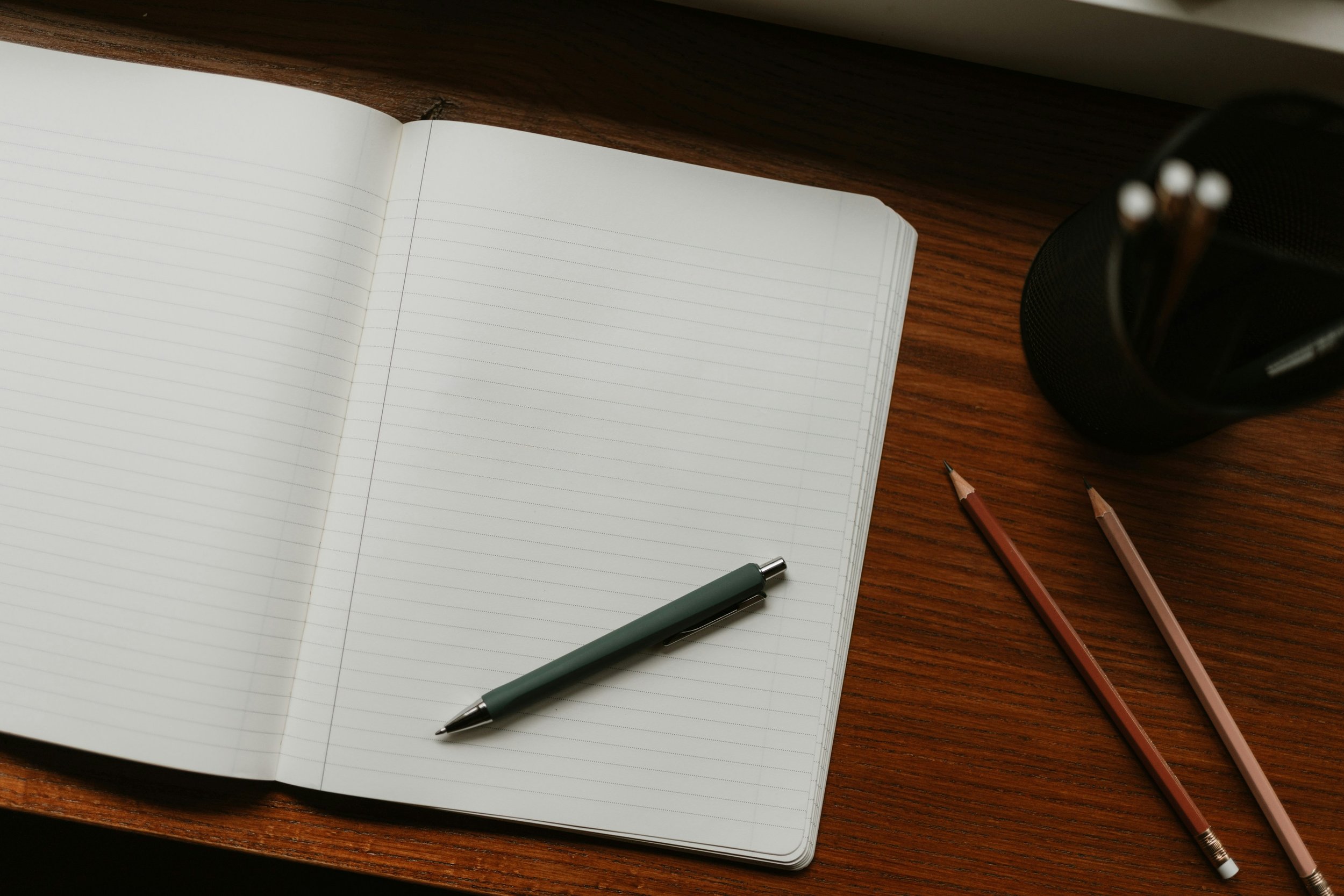 Open lined notebook on wooden desk with three colored pencils, a pen, and a black pen holder containing more pens.
