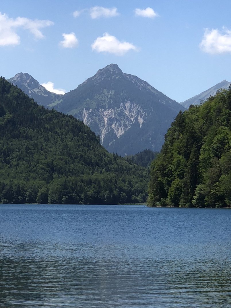 A serene mountain landscape with a calm lake in the foreground, green forested hills on either side, and tall mountains with rocky peaks in the background under a partly cloudy blue sky.