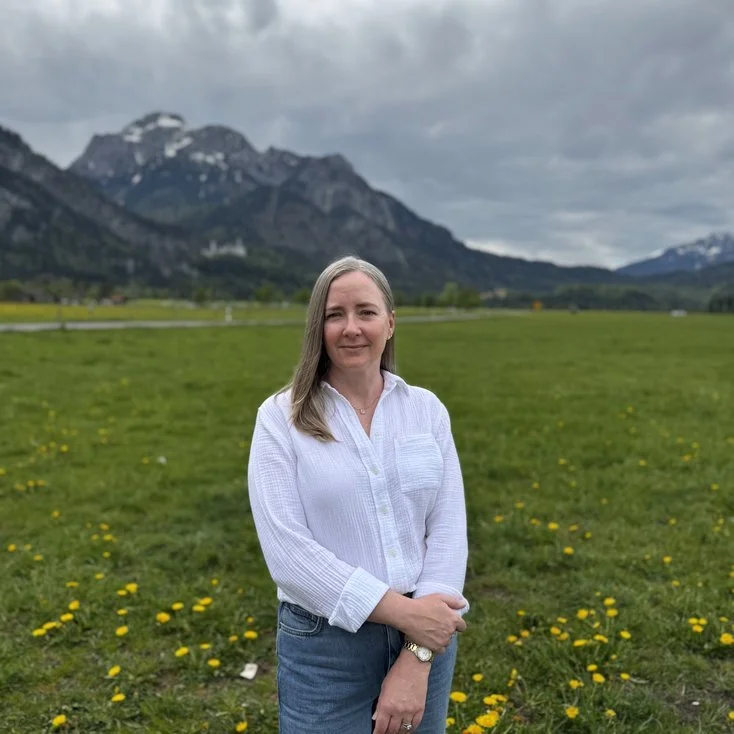 A woman in a white blouse and jeans standing in a green field with yellow flowers, mountains in the background, and a cloudy sky.