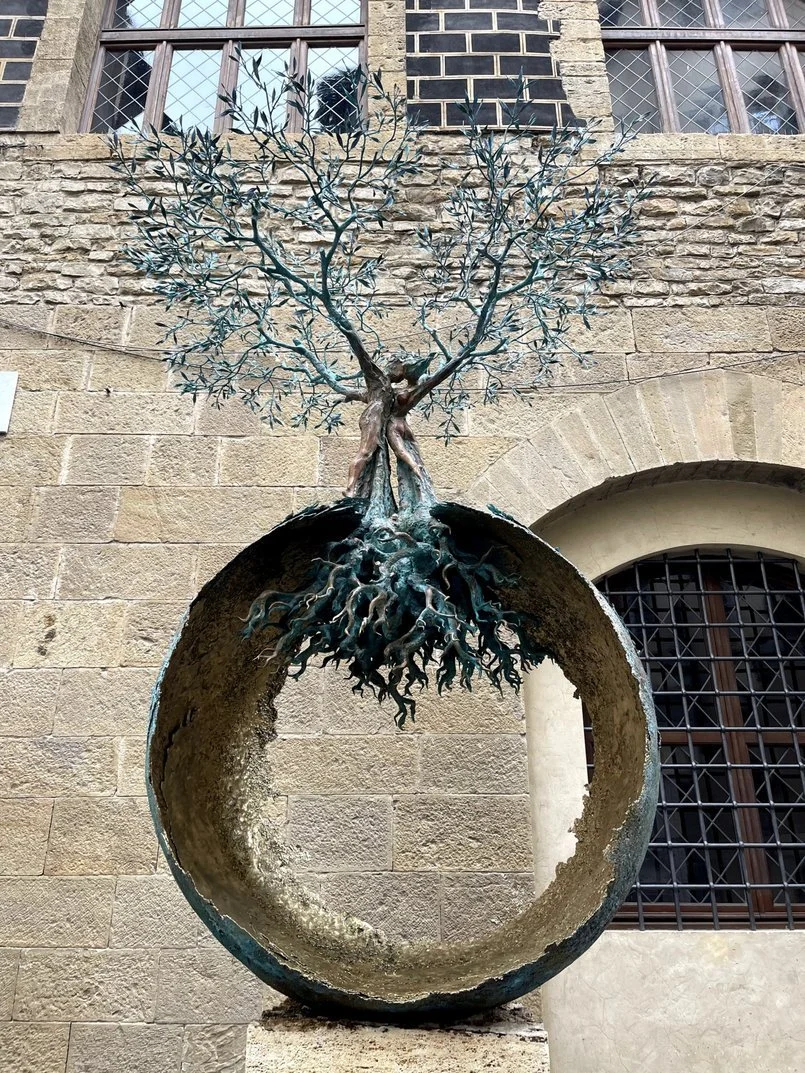 Bronze sculpture of a tree with roots inside a hollow circular base, placed outdoors against stone walls with windows.