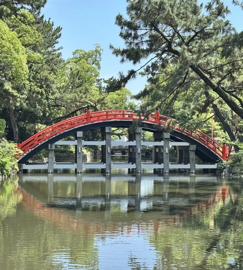 A red arched bridge over a body of water surrounded by green trees and blue sky.