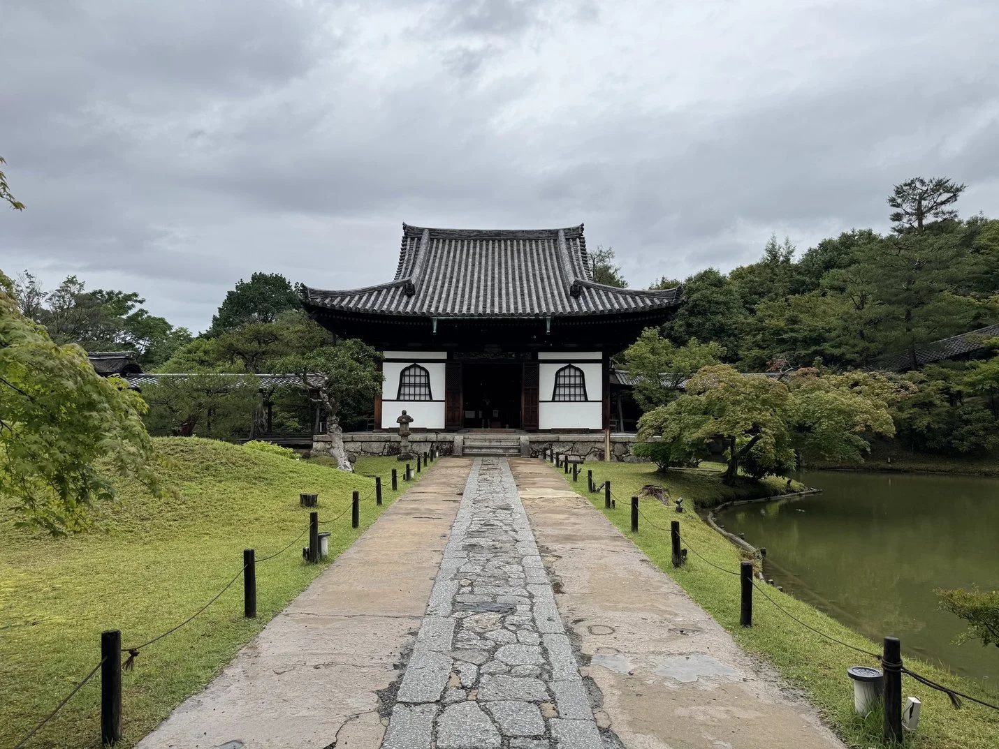 A traditional Japanese building with a curved roof, surrounded by trees and a pond, with a stone pathway leading to the entrance.