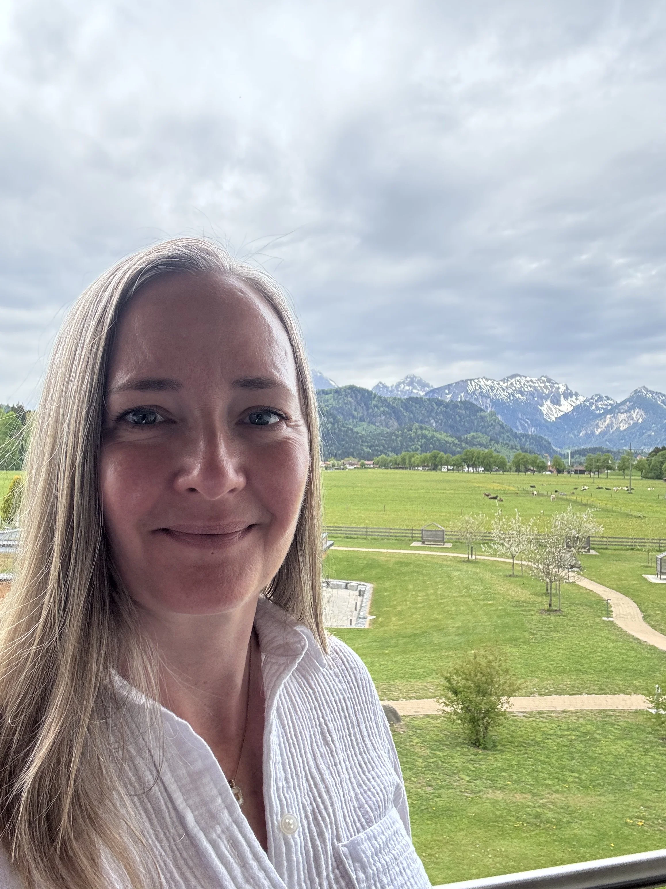 Close-up of a woman with blonde hair smiling outdoors, with mountains, cloudy sky, lush greenery, and a field with sheep in the background.