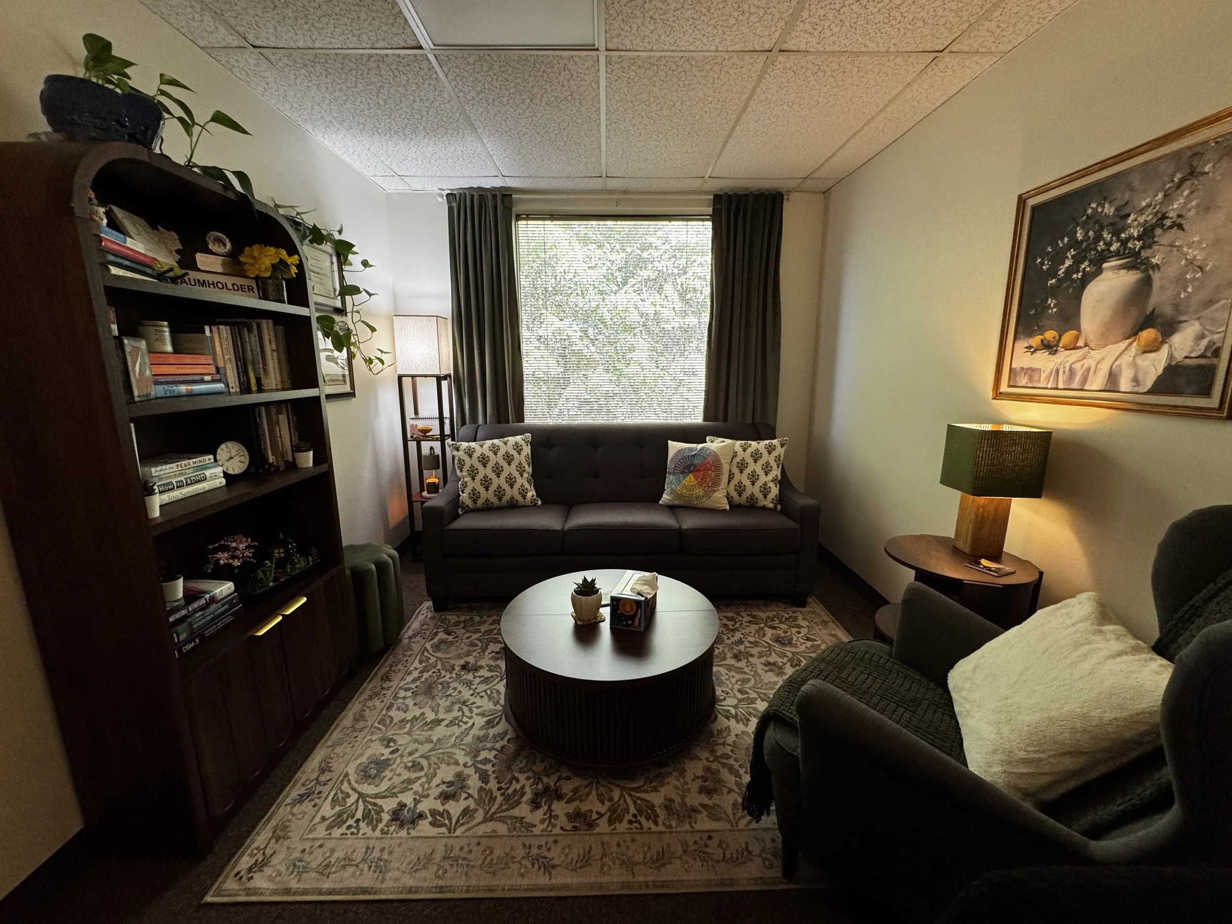 A cozy living room with dark brown sofa chairs, a wooden coffee table, a tall bookshelf, picture frames, potted plants, a painting of flowers in a vase, and a window with dark curtains letting in natural light.
