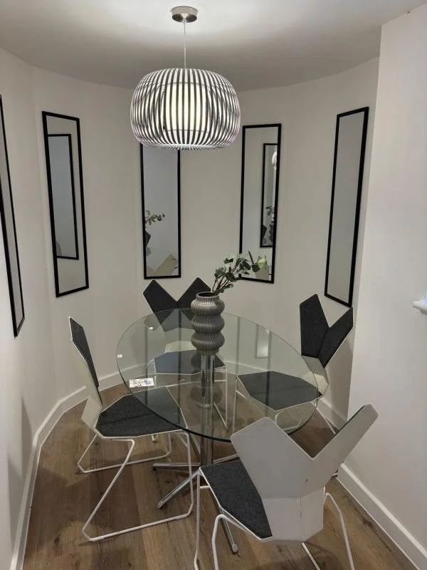 Modern dining room with a glass oval table, six black and white chairs, a large pendant light, and framed mirrors on the white walls with hardwood flooring.