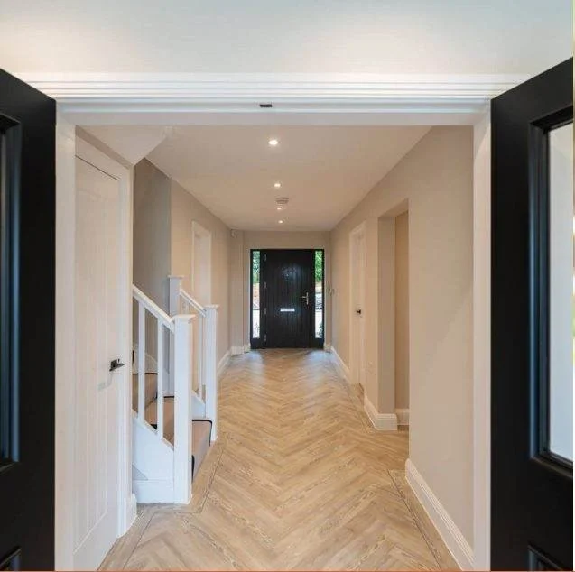 View of a modern hallway with light-colored walls, wooden herringbone floor, and a black front door with glass panels, seen from an interior doorway.