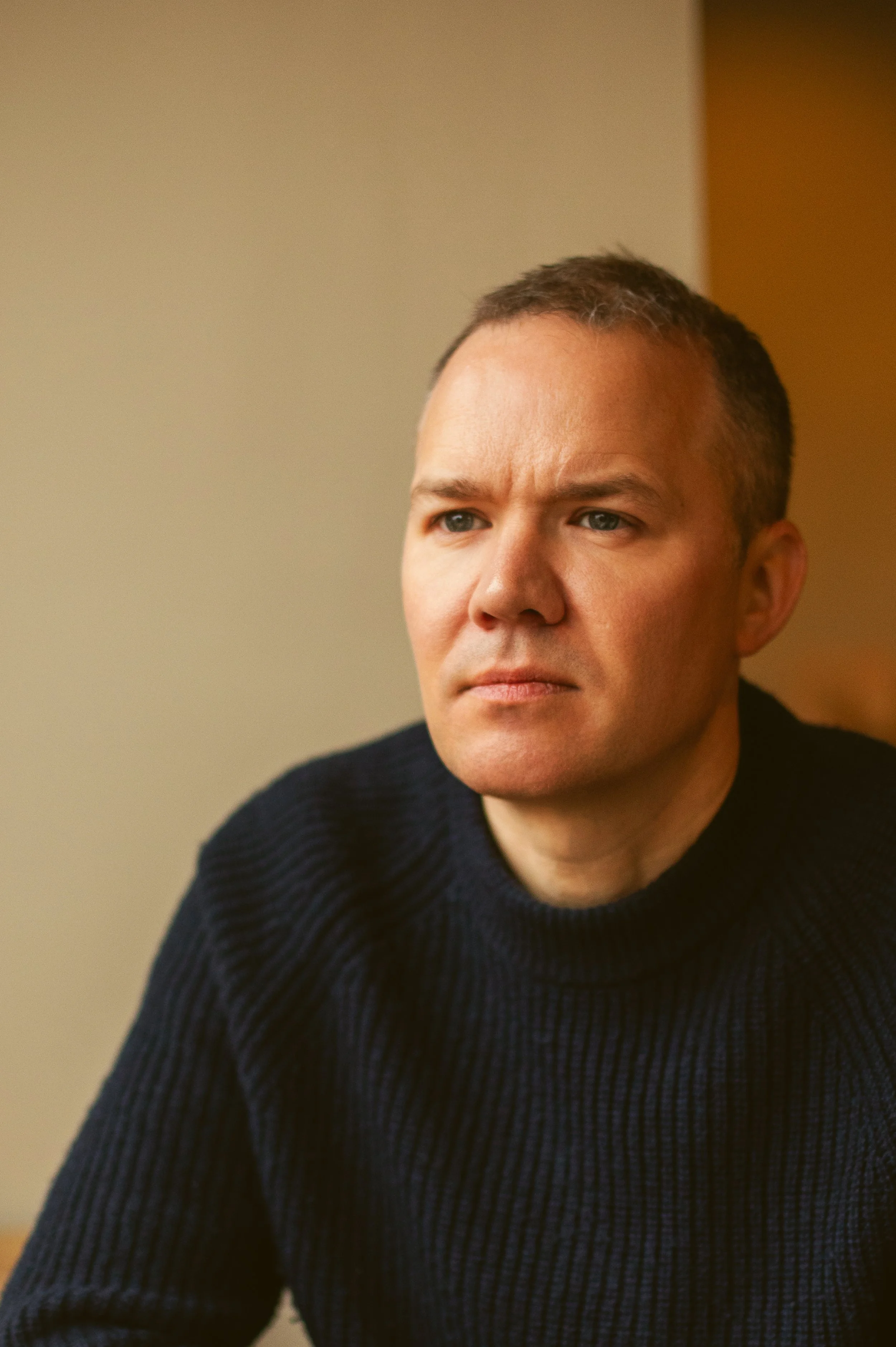 A man with short brown hair and blue eyes, wearing a dark navy blue ribbed sweater, sitting indoors with a neutral expression.
